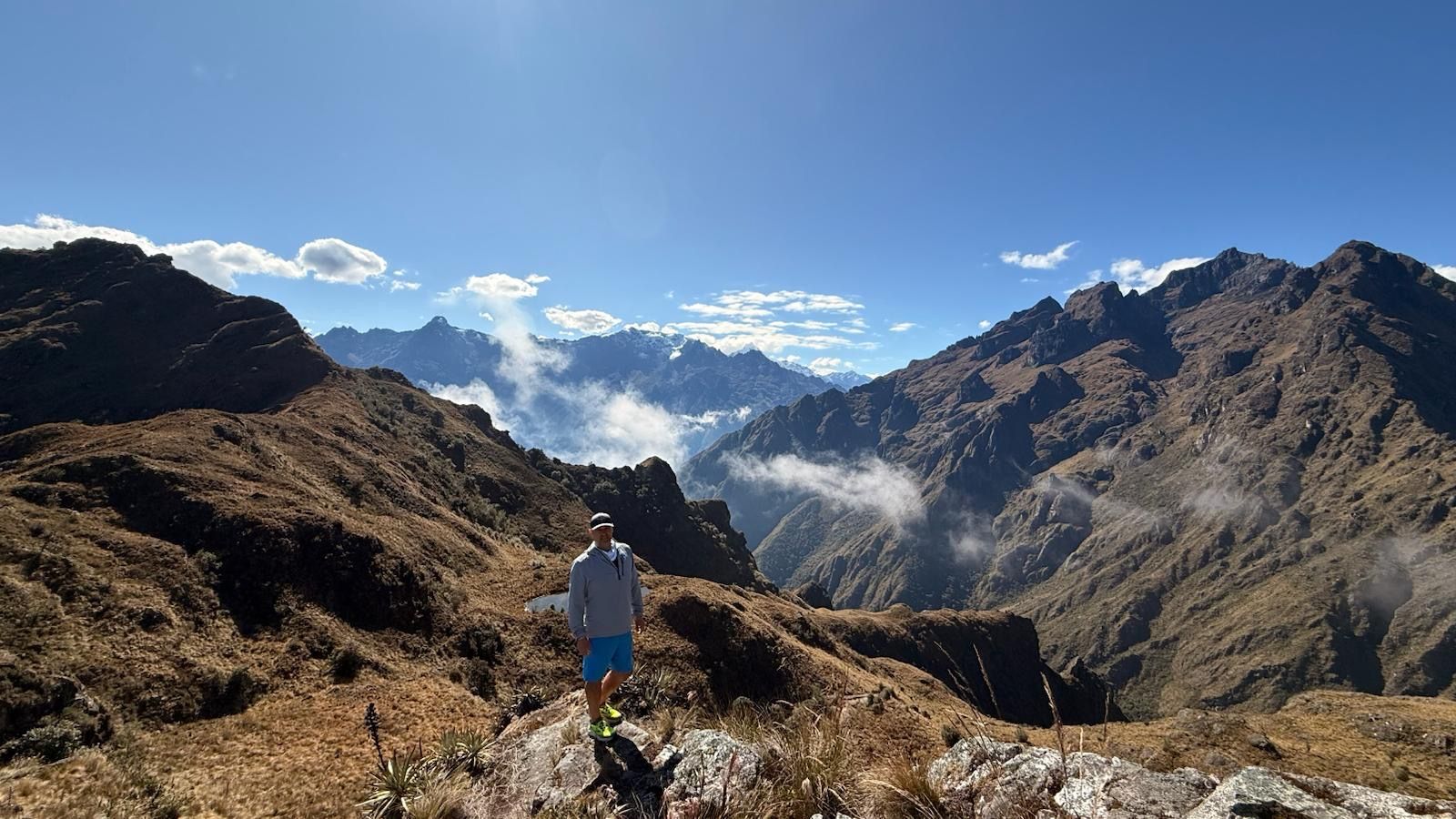 Man on a mountaintop, overlooking a valley with clouds and mountains. Sunny, blue sky.
