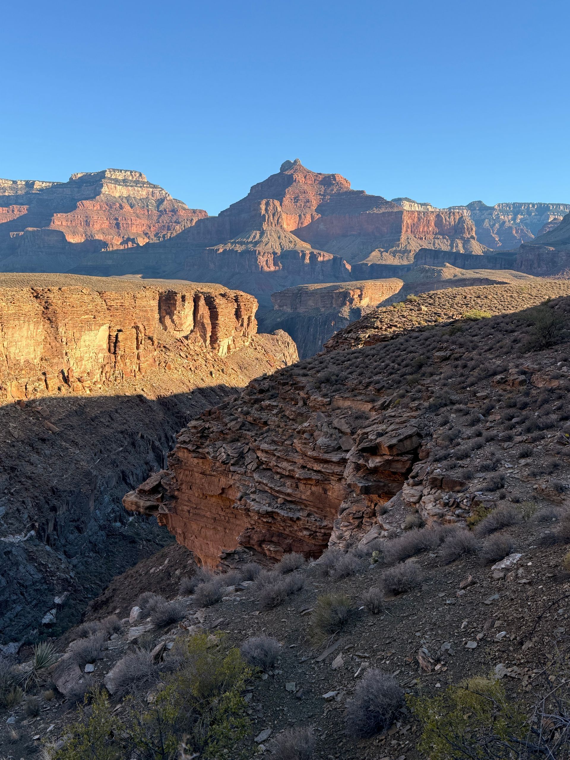 Canyon landscape with red rock formations under a clear blue sky. Shadows cast on the rocky terrain.