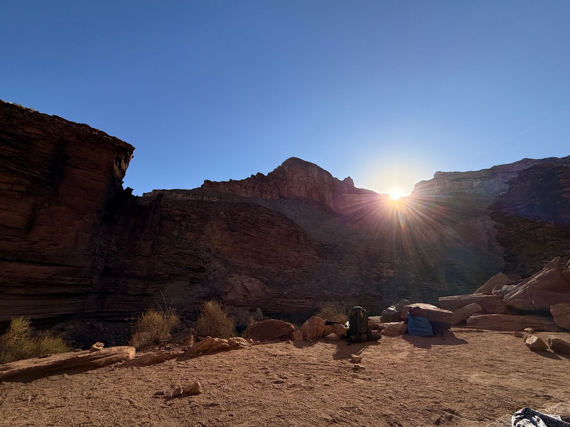 Sun shining over a canyon with brown rock formations. A person sits with a blue backpack in the foreground.