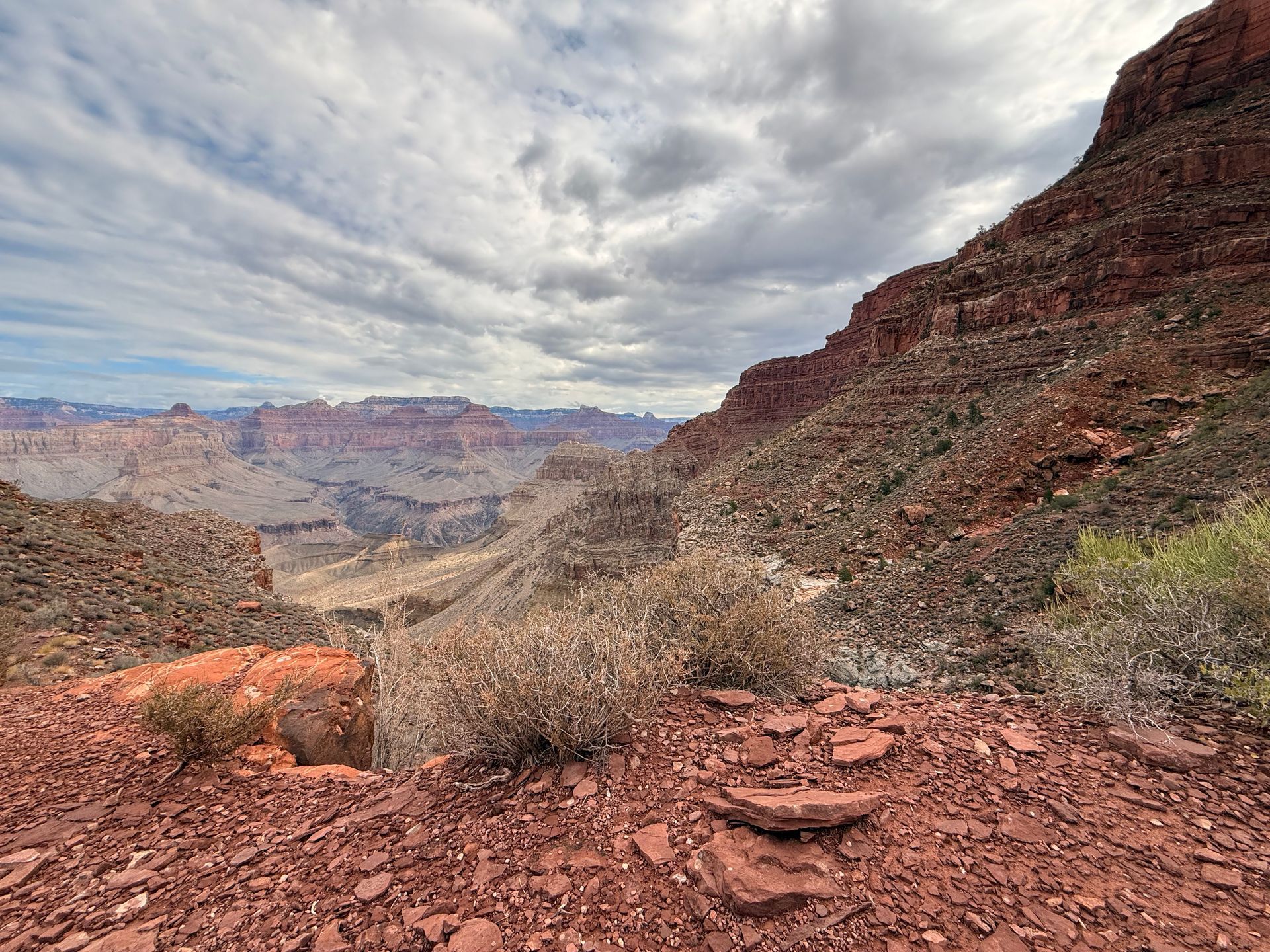 Canyon landscape with red rock formations, cloudy sky, and sparse vegetation.