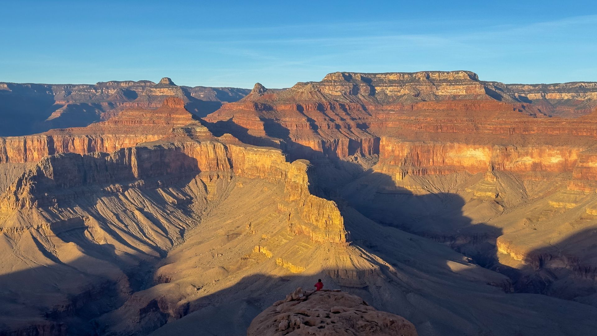 Grand Canyon vista at sunrise, warm colors illuminate rock formations; a person sits on a rock outcrop.