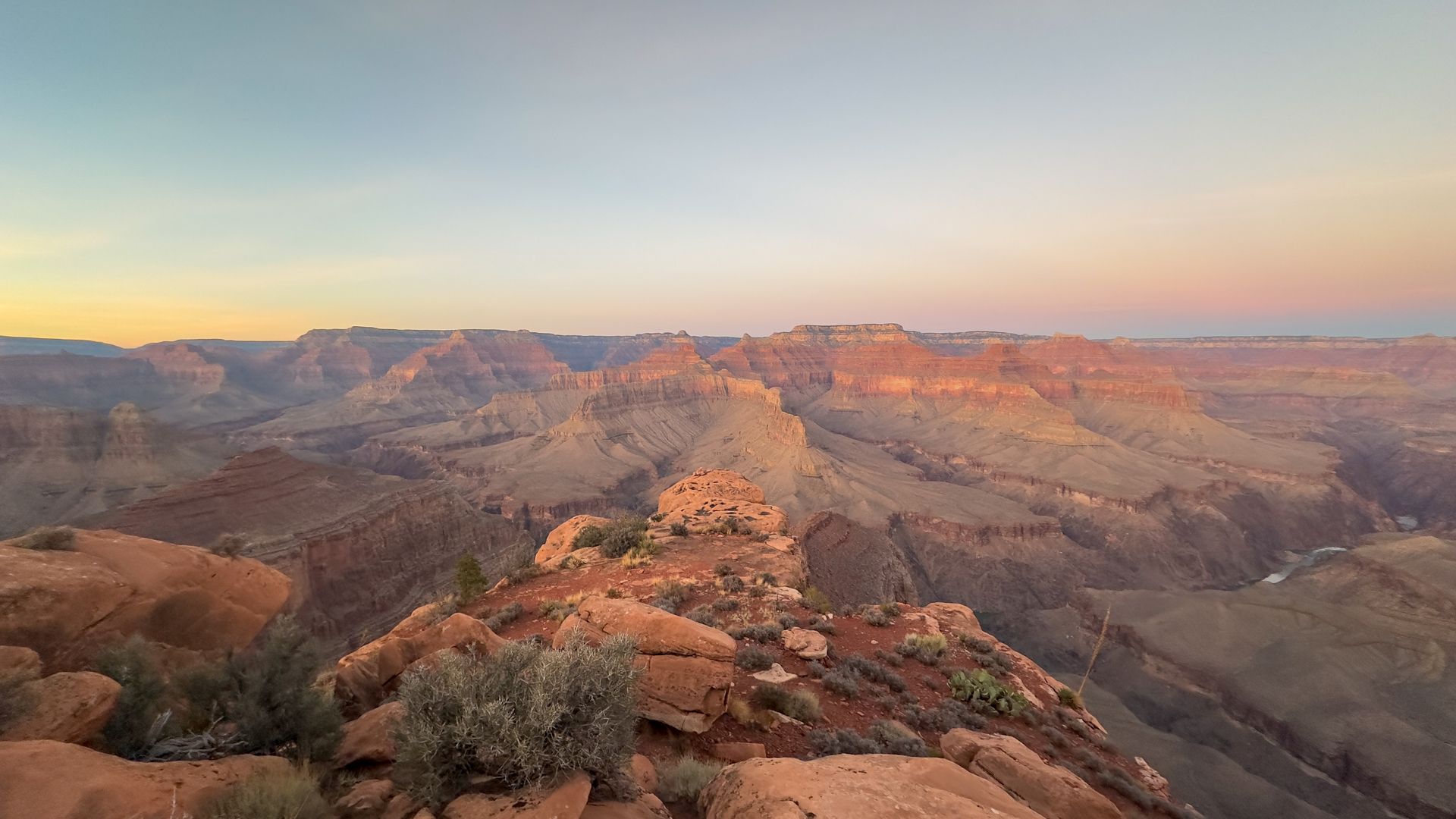 Panoramic view of the Grand Canyon at sunrise; layers of canyons, in shades of orange and brown, under a pink and blue sky.