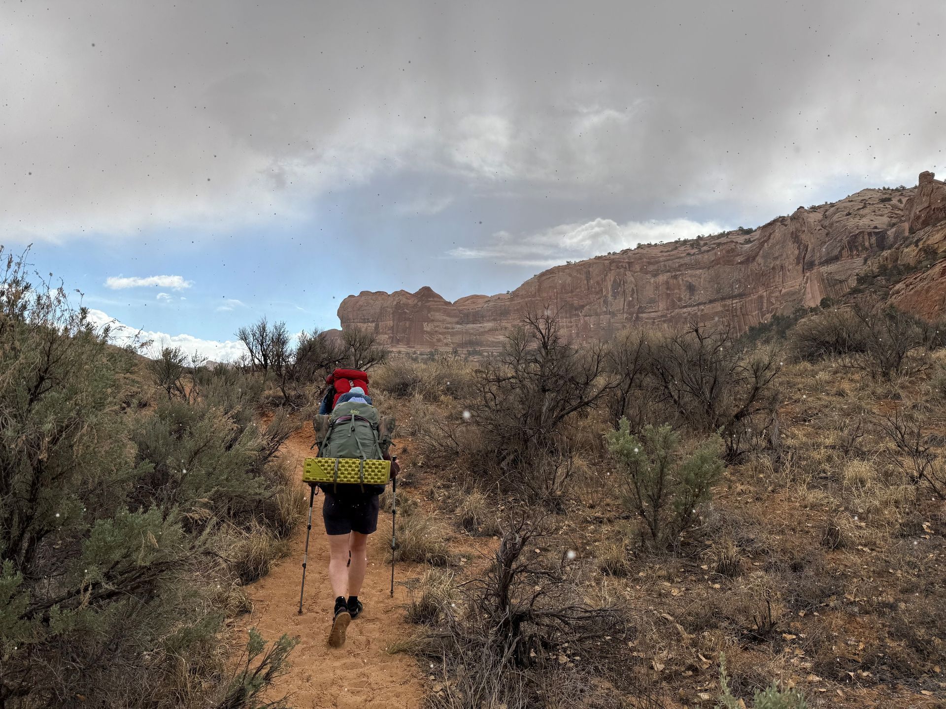 Person hiking on a dirt path in a desert landscape with backpack and trekking poles, under a cloudy sky.