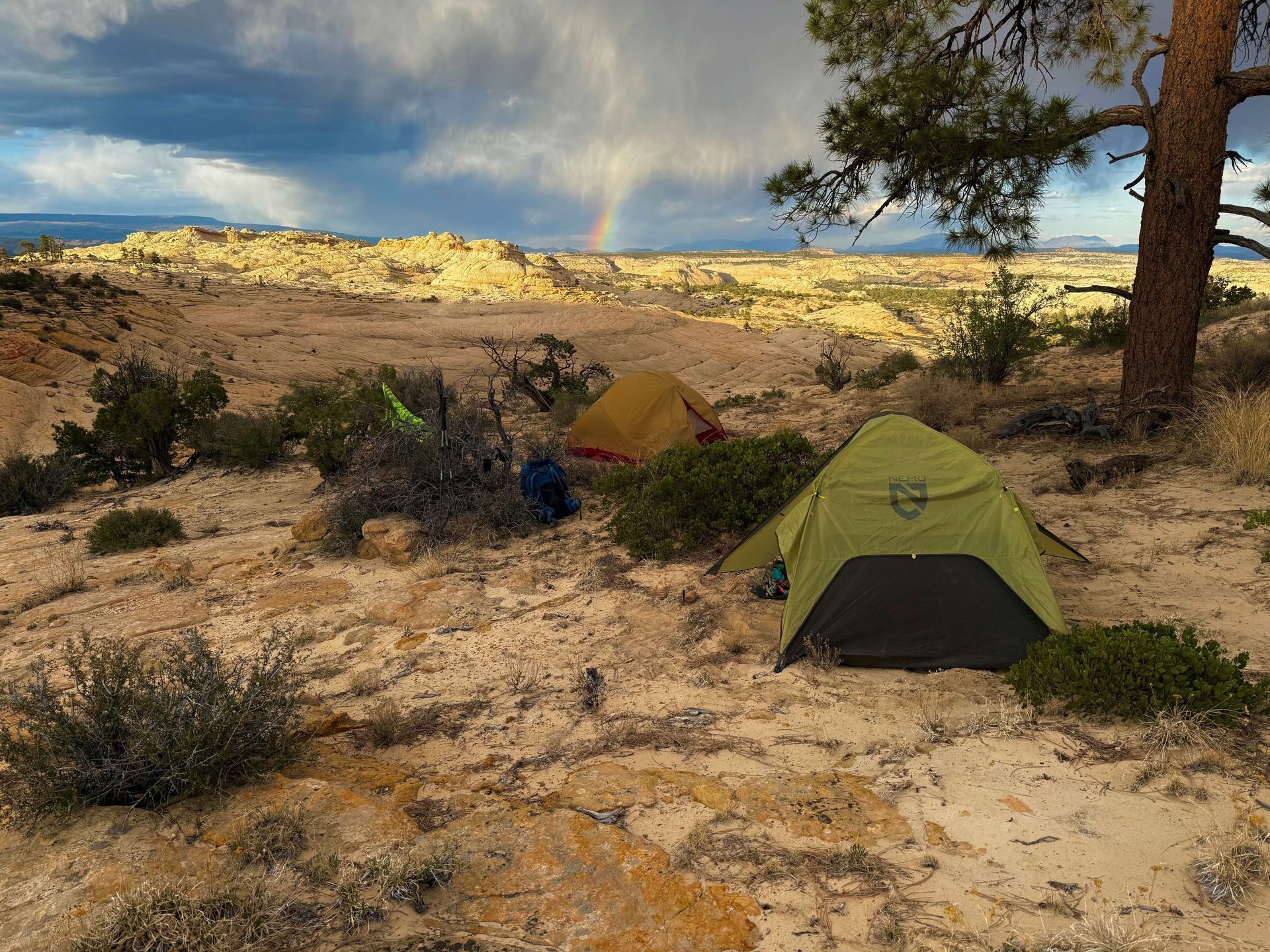 Two tents are pitched on a desert plateau, with a rainbow in the background under a cloudy sky.