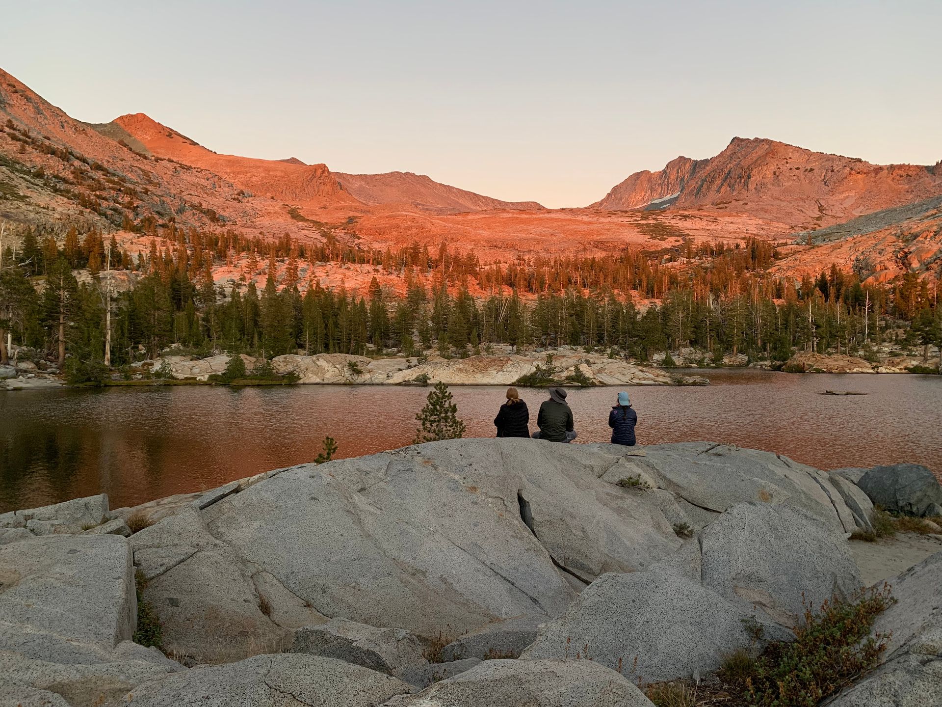 Three people are sitting on a rock overlooking a lake with mountains in the background.