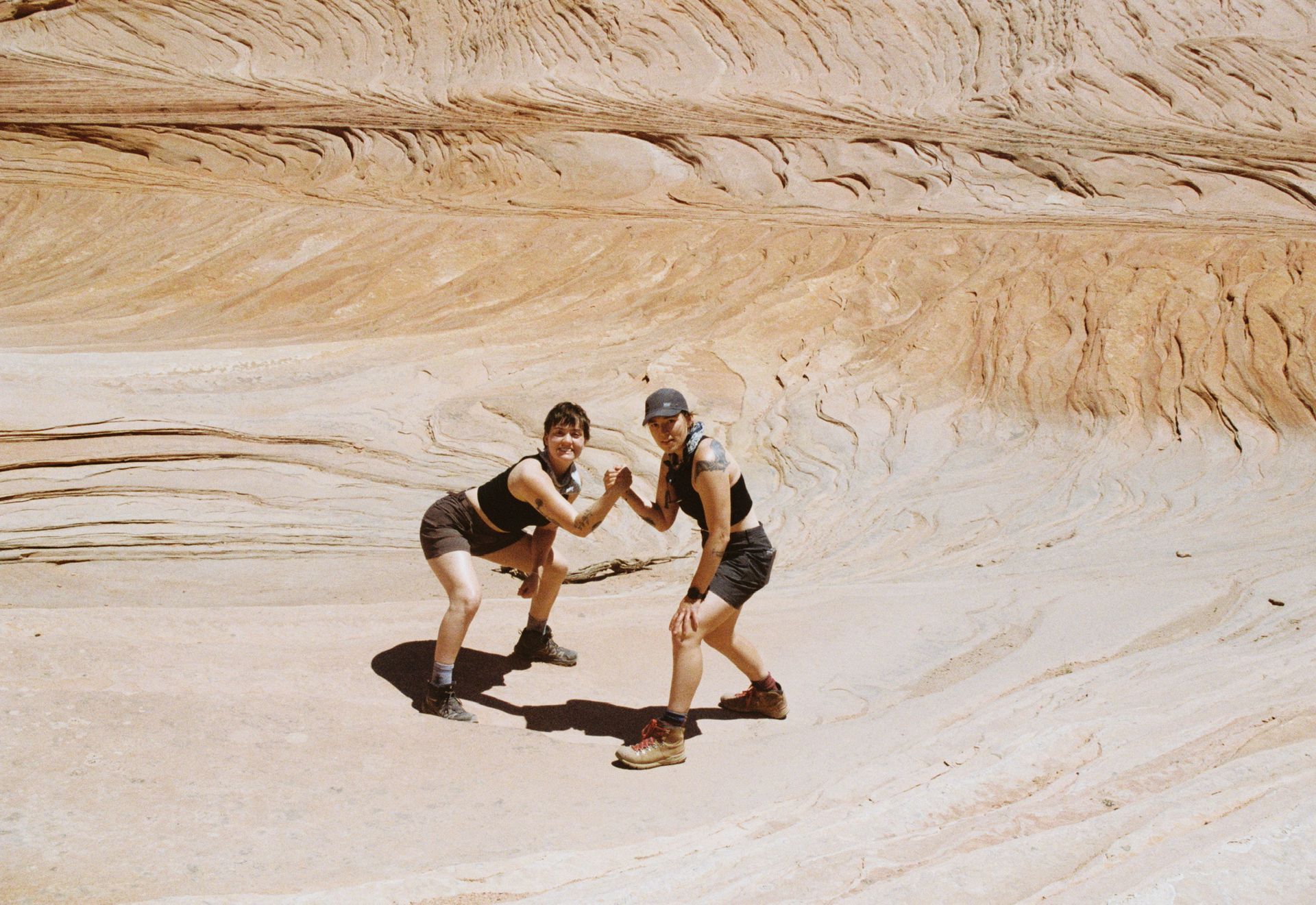 Two people doing a fist bump in a canyon. They wear shorts, tanks, and boots. Sand-colored canyon walls.
