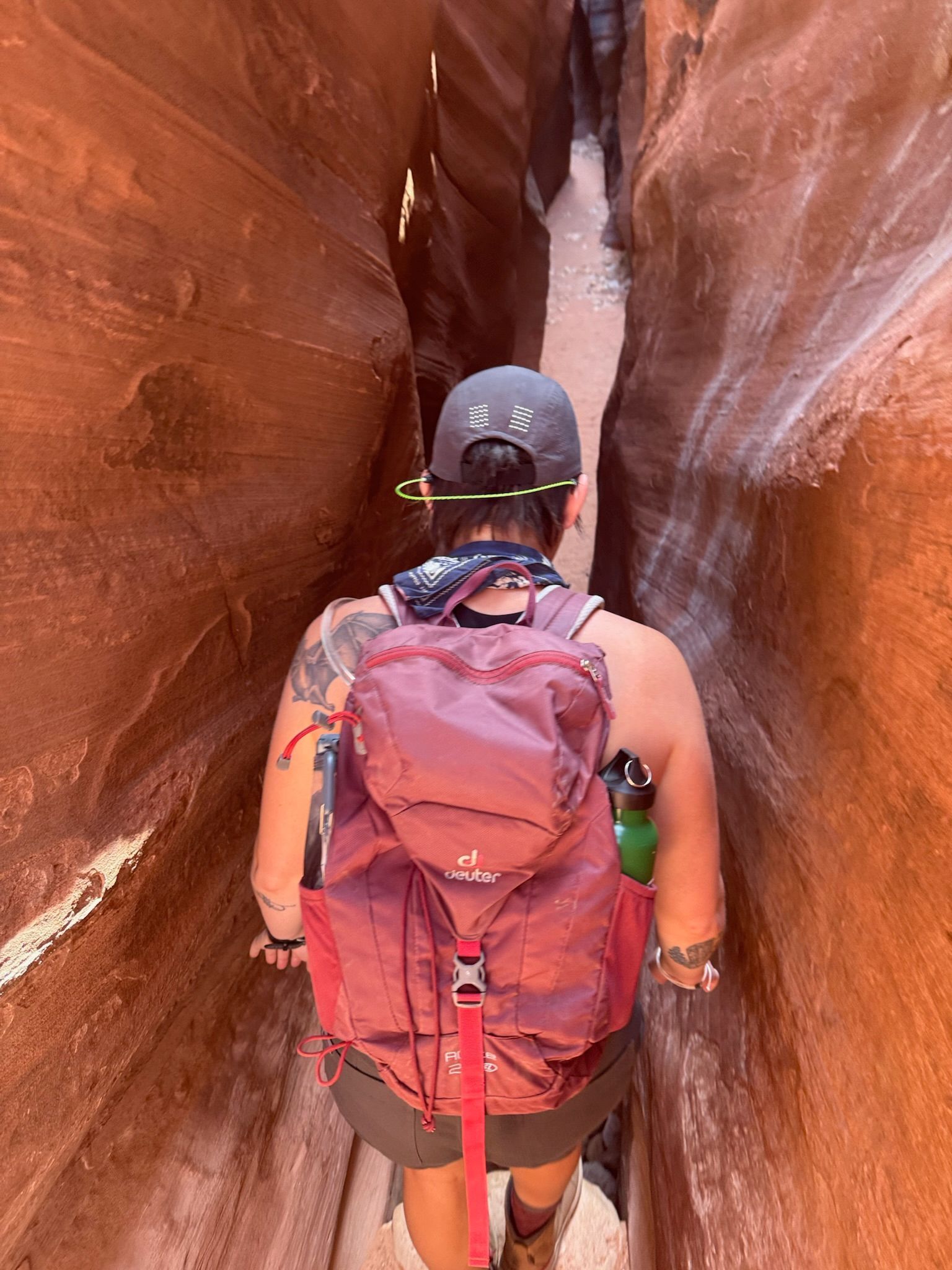 Person hiking in a narrow slot canyon, wearing a backpack, between red rock walls.