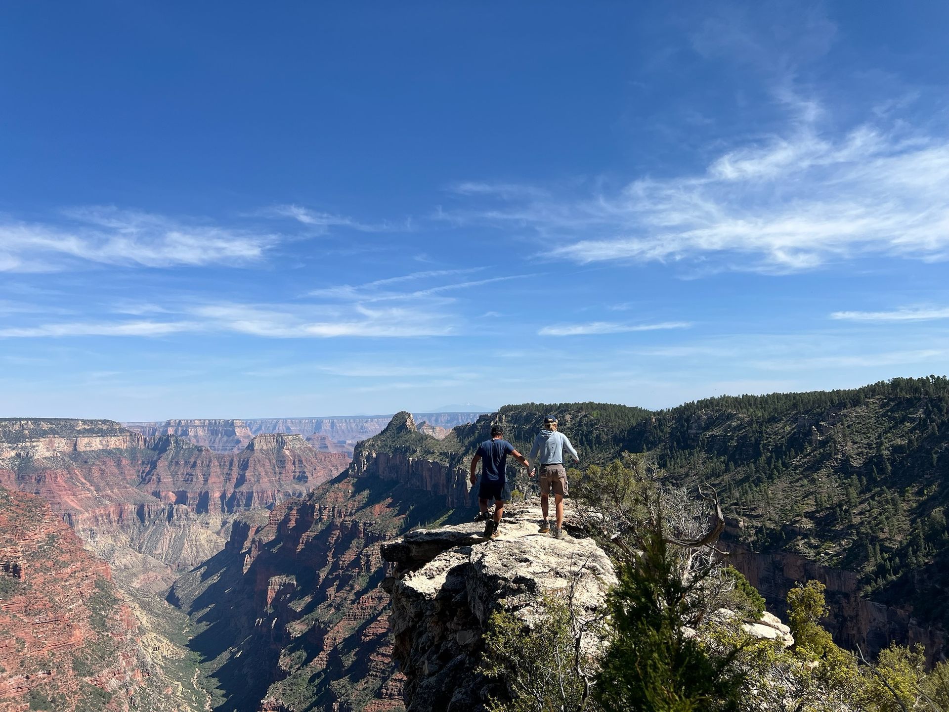 Two people stand on a cliff, overlooking a canyon under a blue sky.