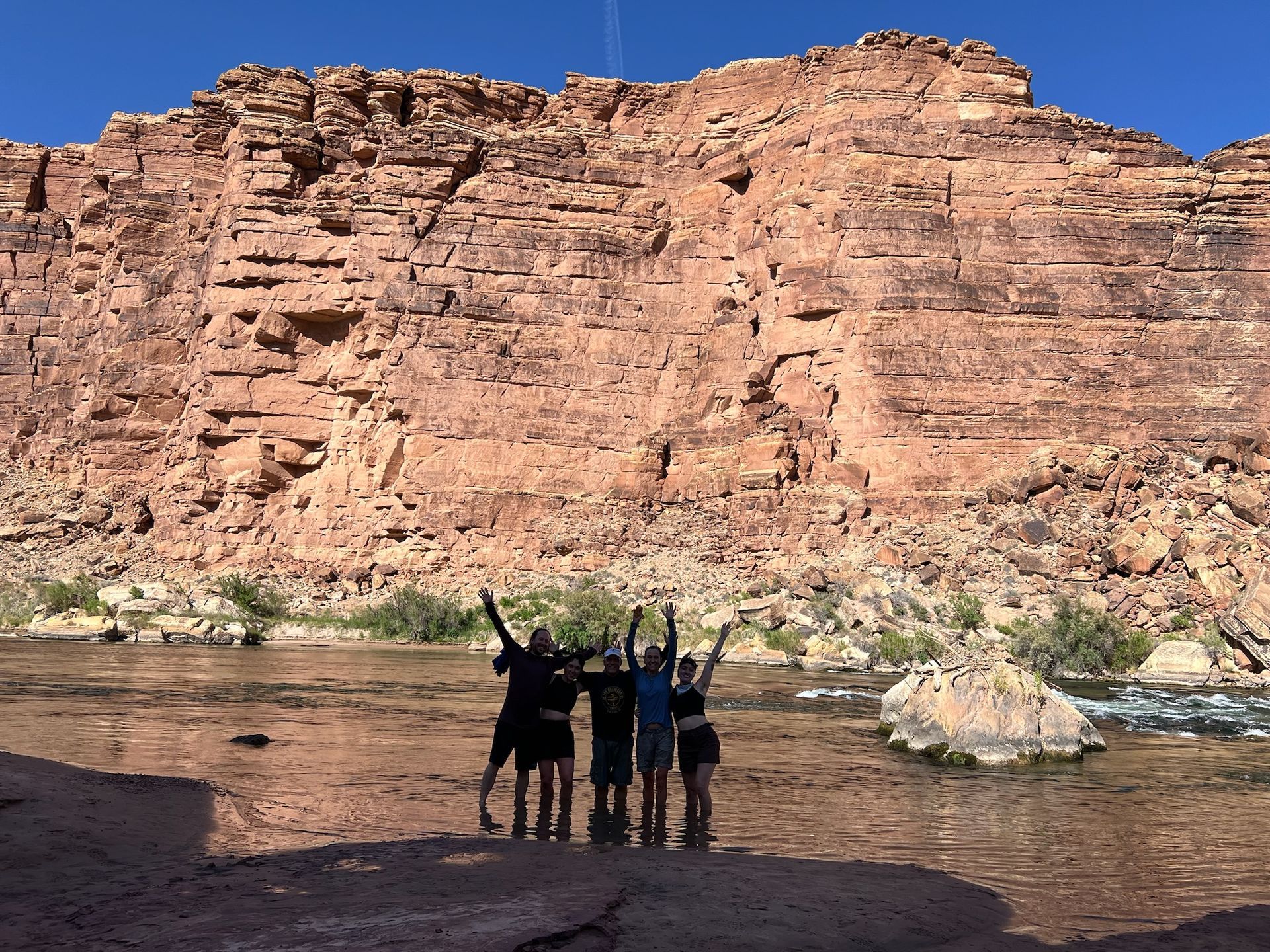 Five people stand with arms raised in front of a canyon wall and a river.