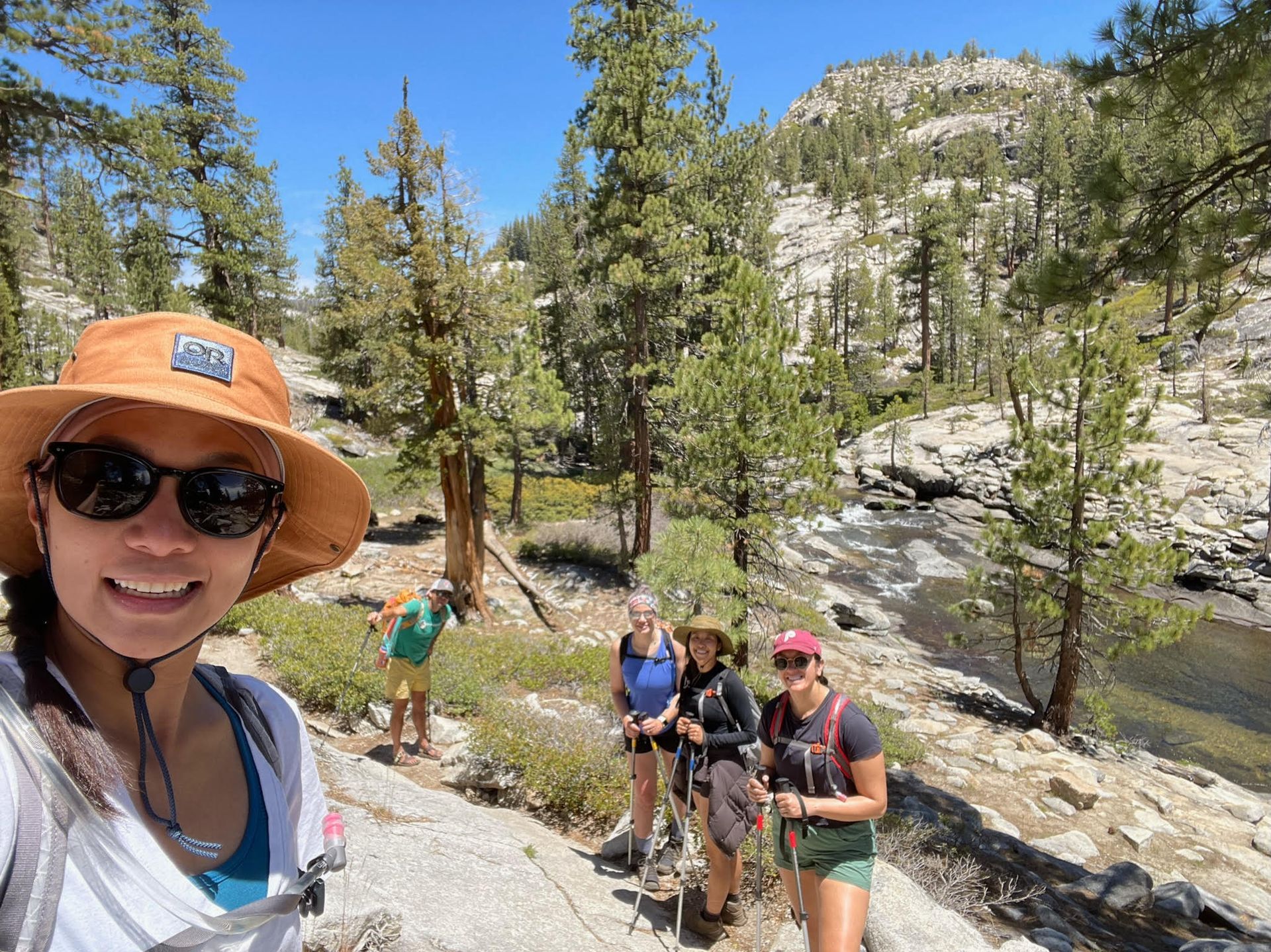 A woman wearing a hat and sunglasses is taking a selfie with her friends while hiking.