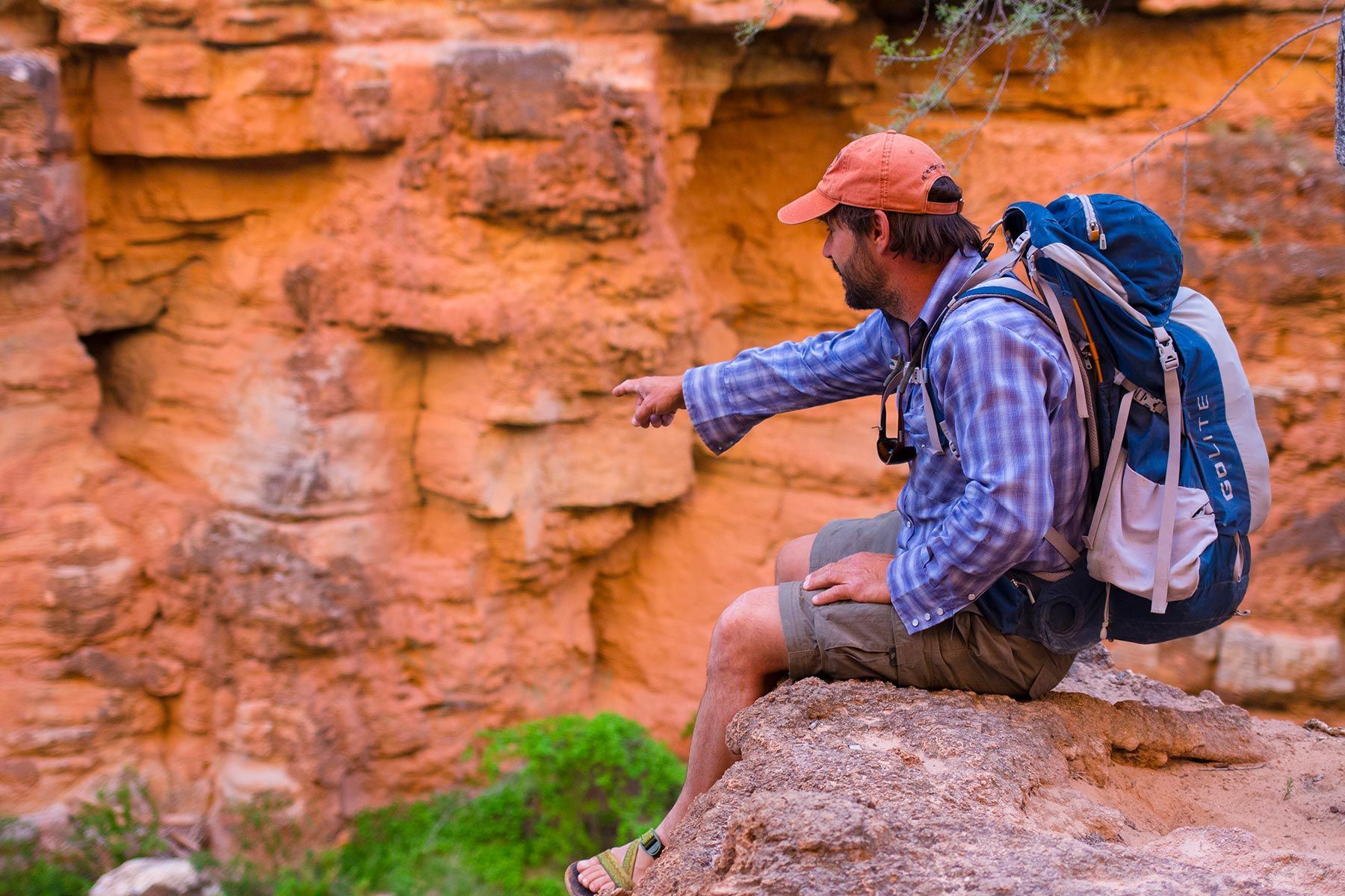 A man with a backpack is sitting on a rock pointing at something.