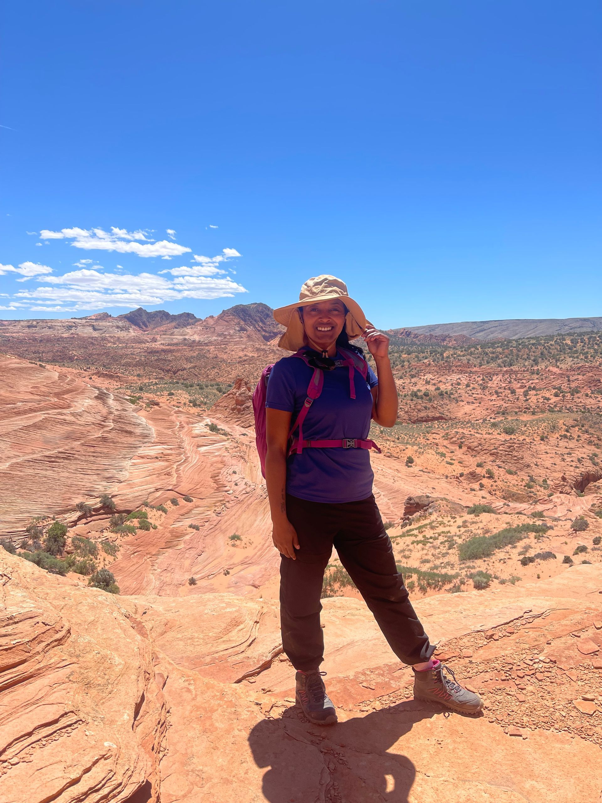 Person with a backpack poses on a red rock formation, blue sky background.