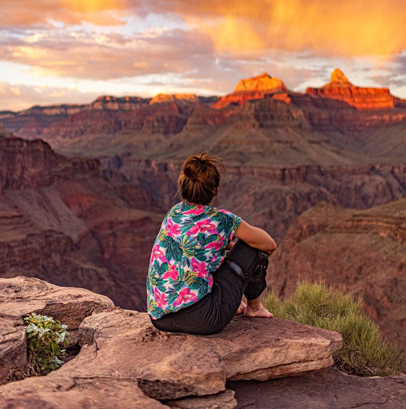 Person sitting on a rock overlooking a canyon at sunset, wearing a floral shirt.