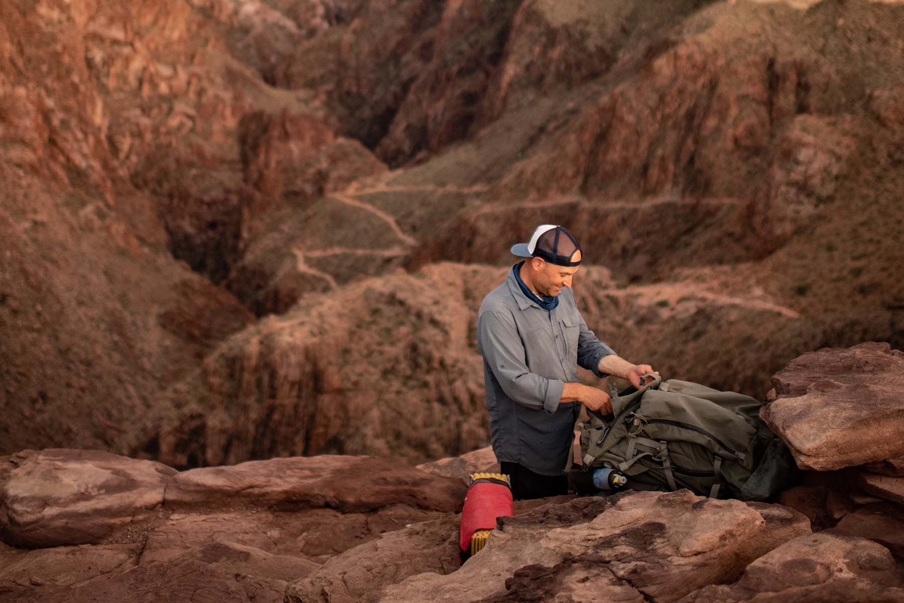 Man in cap, packs backpack on red rock ledge, canyon backdrop.