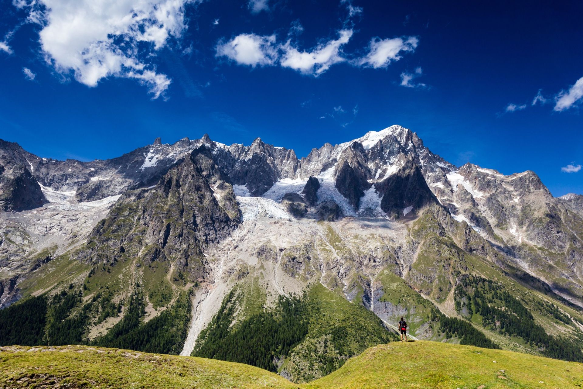 Mountain range with snow-capped peaks under a blue sky with clouds; green hillside and one person visible.
