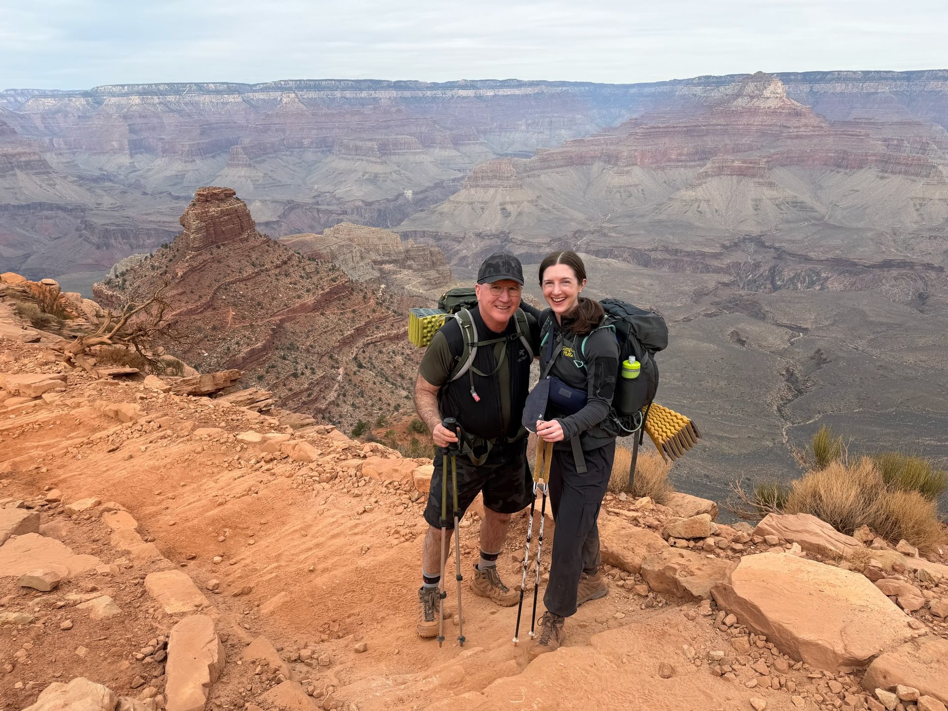 Two hikers with backpacks stand on a canyon rim. They smile at the camera, posing with walking sticks, the vast canyon in the background.