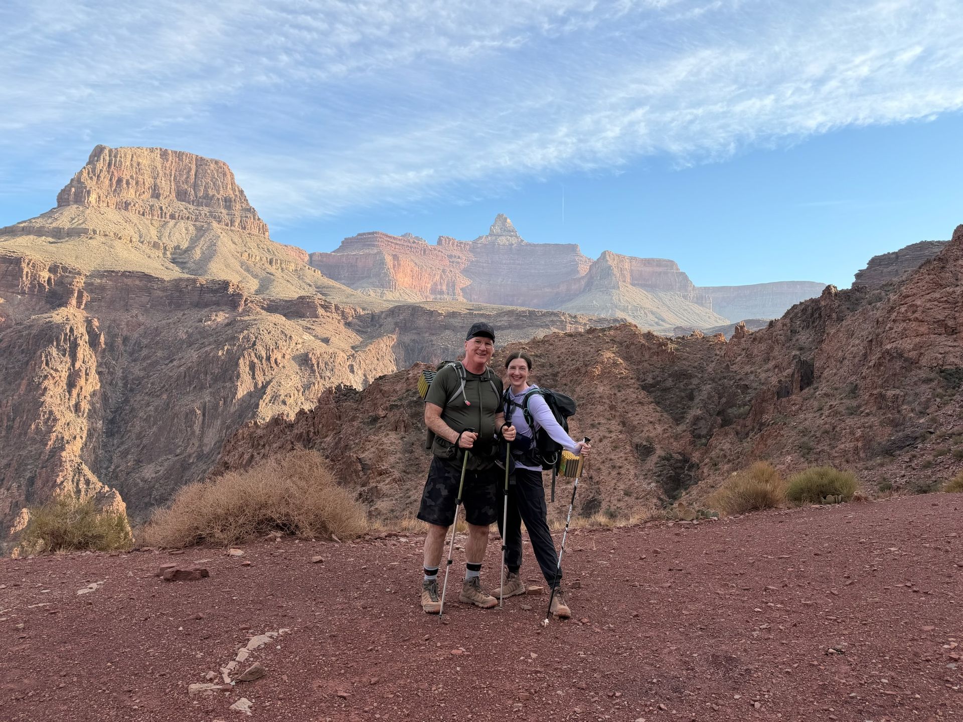 Two hikers pose with the Grand Canyon in the background, blue sky overhead.