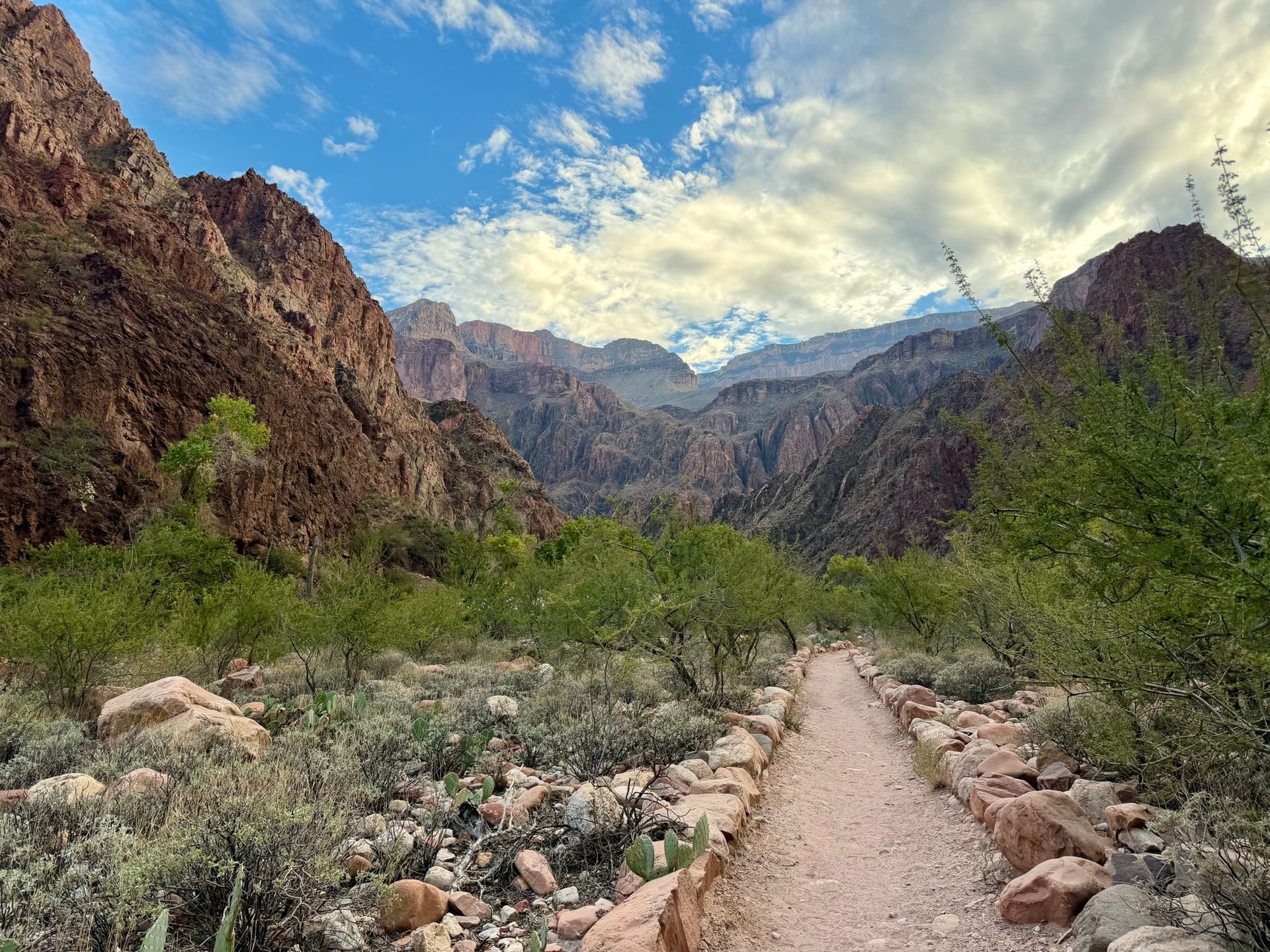 Dirt path winds through a canyon, with reddish-brown rock walls, green bushes, and a cloudy sky.