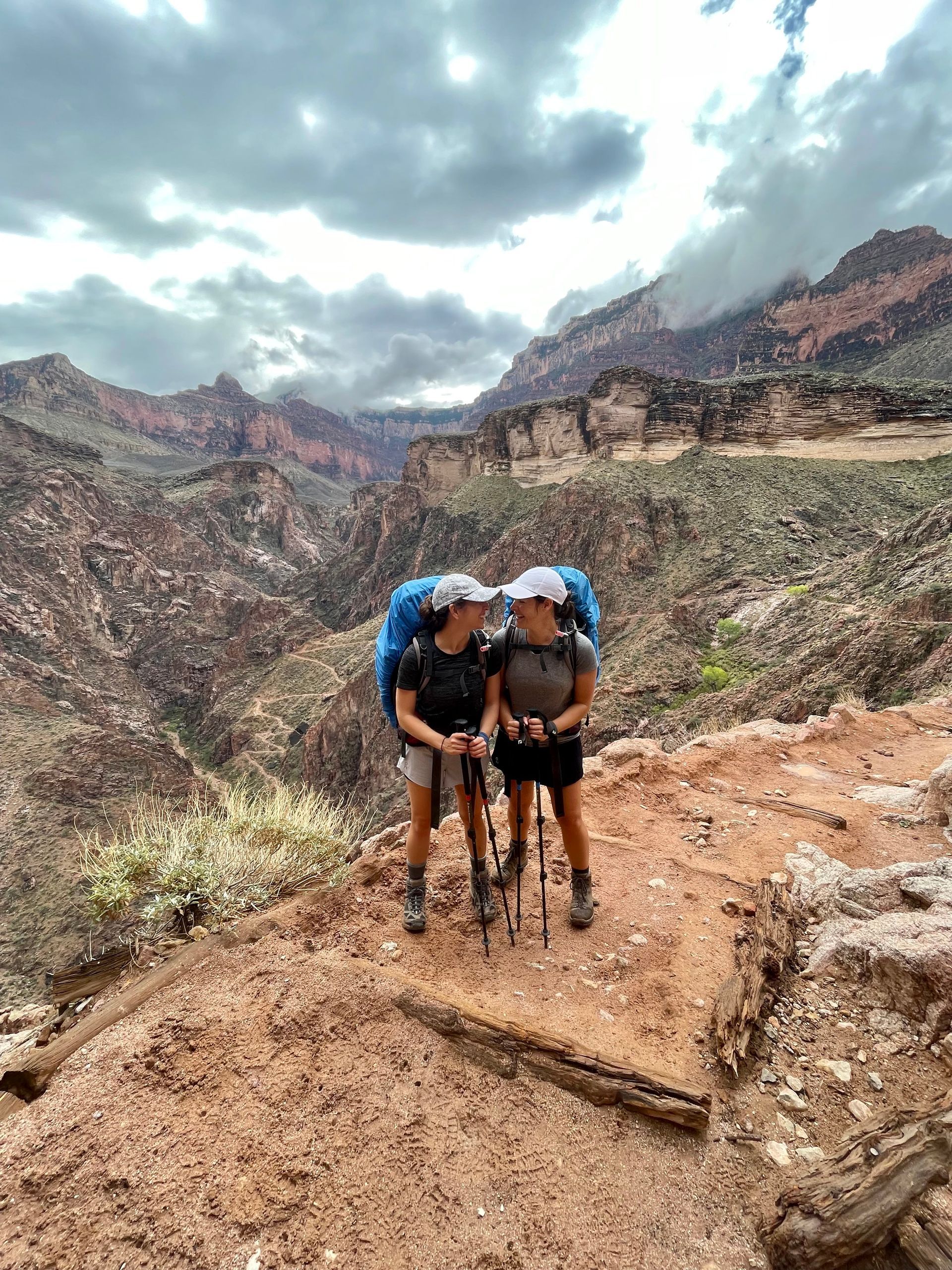 Two women are standing next to each other on a trail in the mountains.