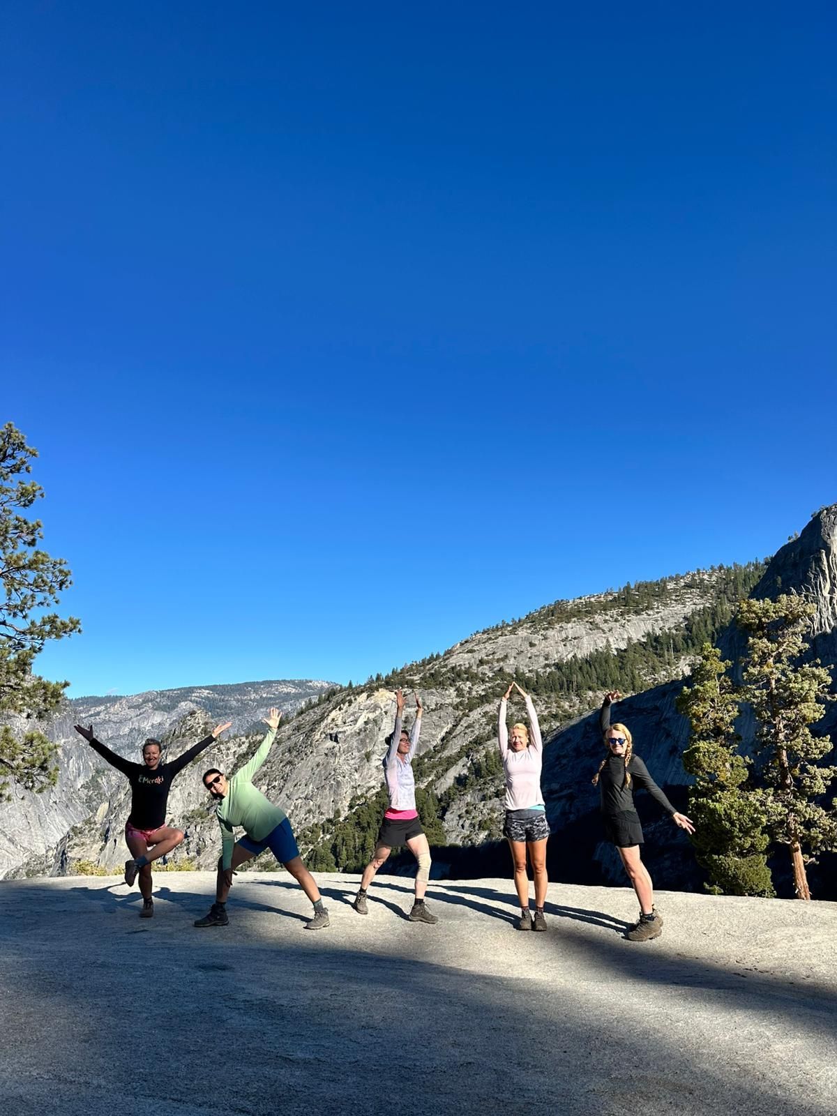 Five people stretching outdoors with arms raised in front of a mountainous landscape. Blue sky.