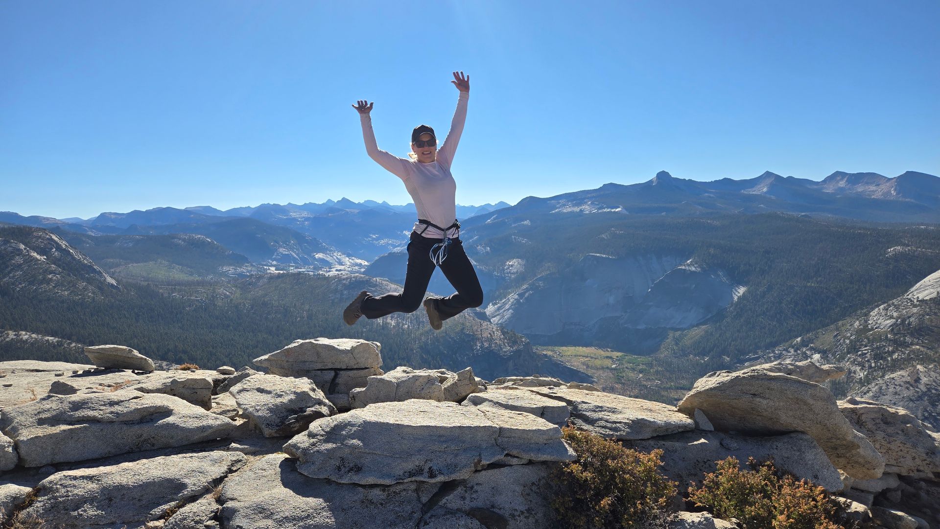 Woman jumps joyfully on a rocky mountain summit, arms raised, with a scenic mountain range in the background under a blue sky.