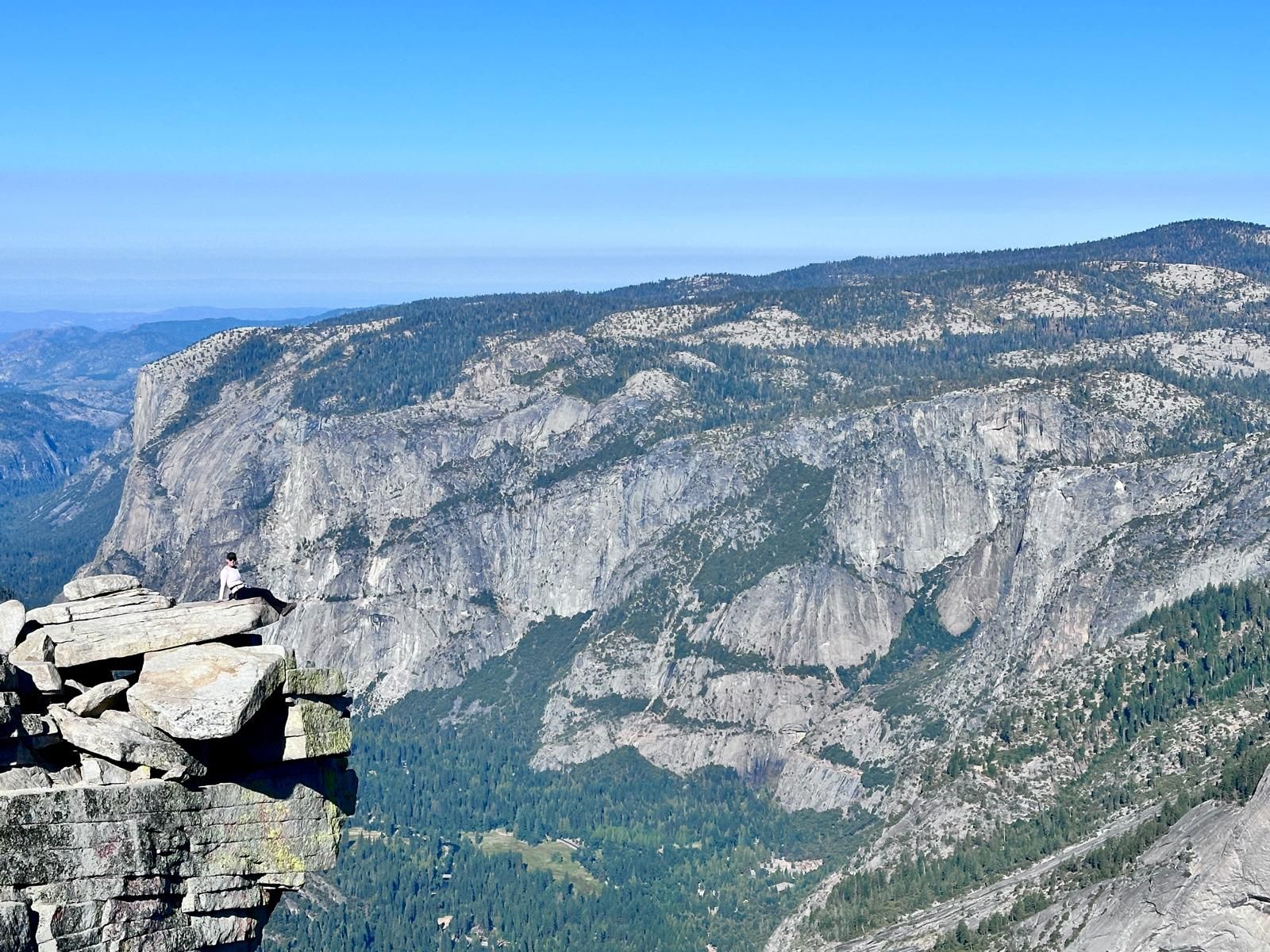 Person sitting on rocky cliff edge, overlooking a vast mountain range with green trees and a blue sky.