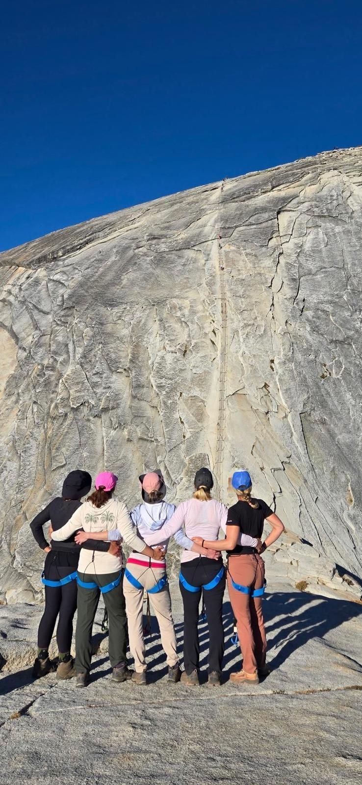 Six people in climbing gear stand arm-in-arm in front of a rock face under a blue sky.