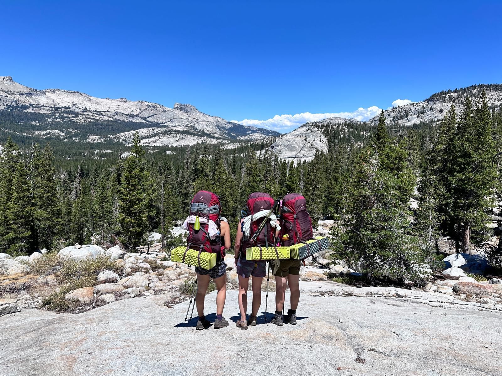 Three backpackers stand on a rocky hilltop, looking at a mountain vista under a blue sky.