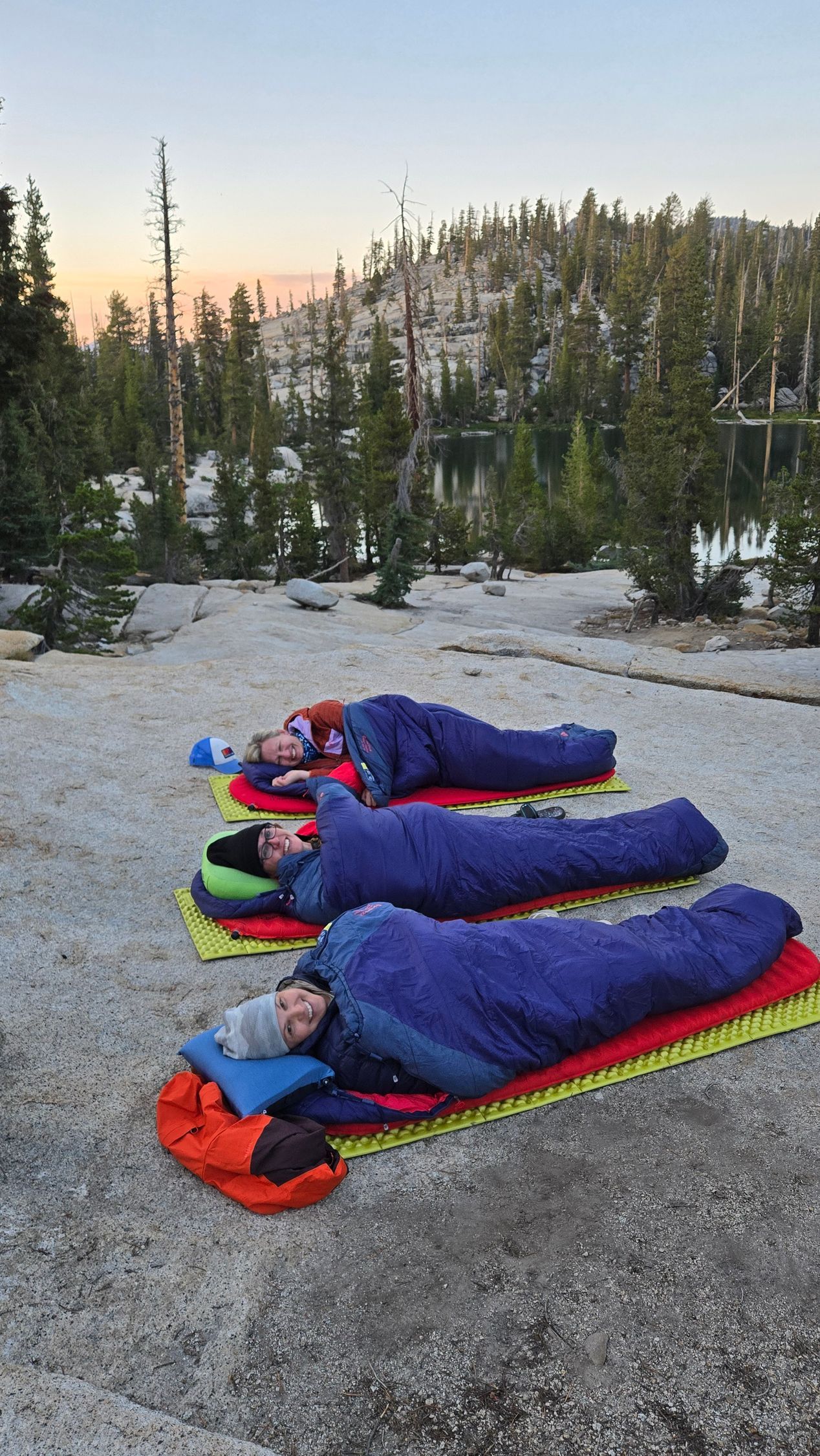 Three people in sleeping bags resting on mats outdoors near a lake and trees.