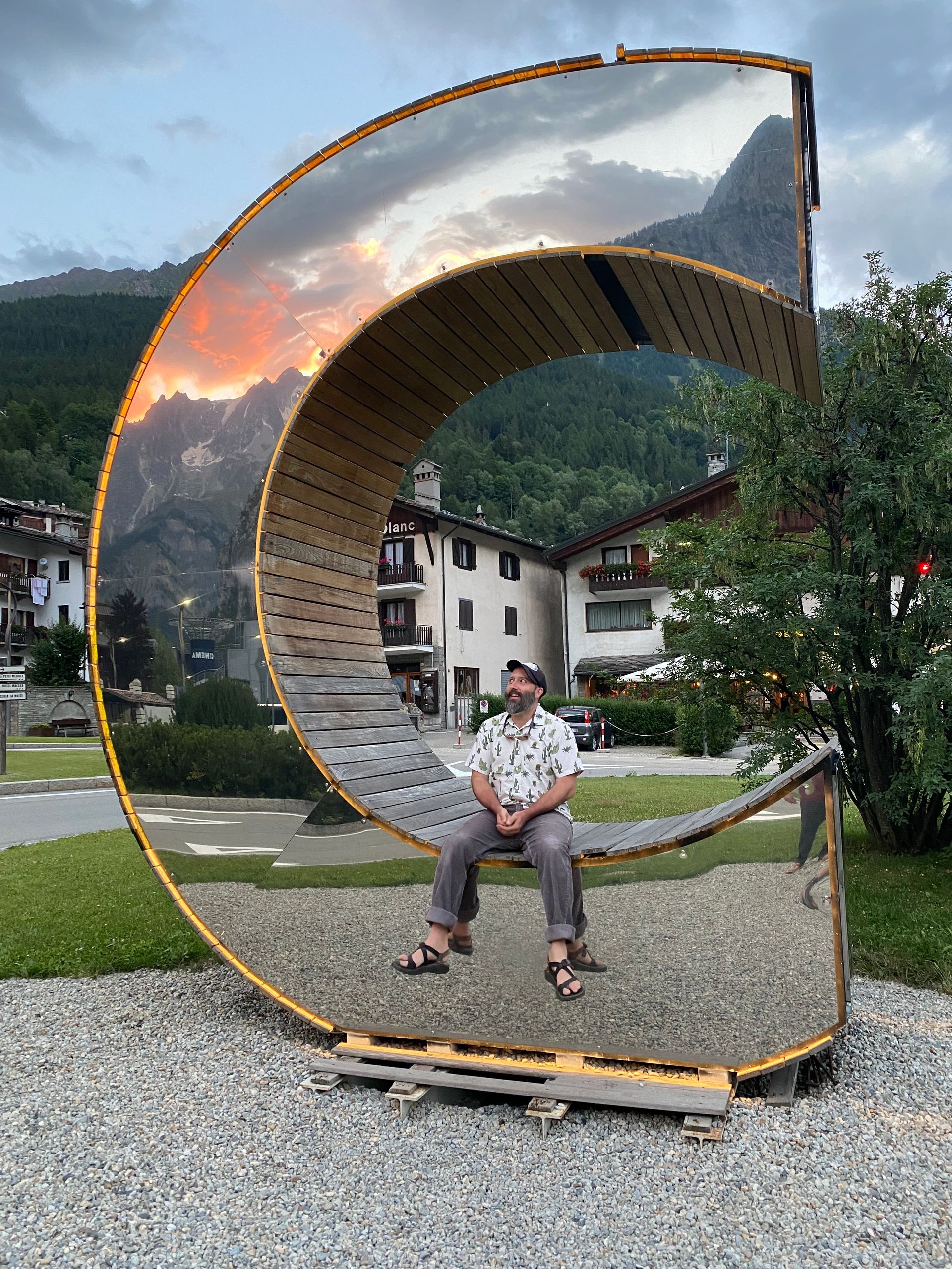 Man seated in front of a mirrored sculpture, reflecting mountains and sunset. Setting is a town with buildings.