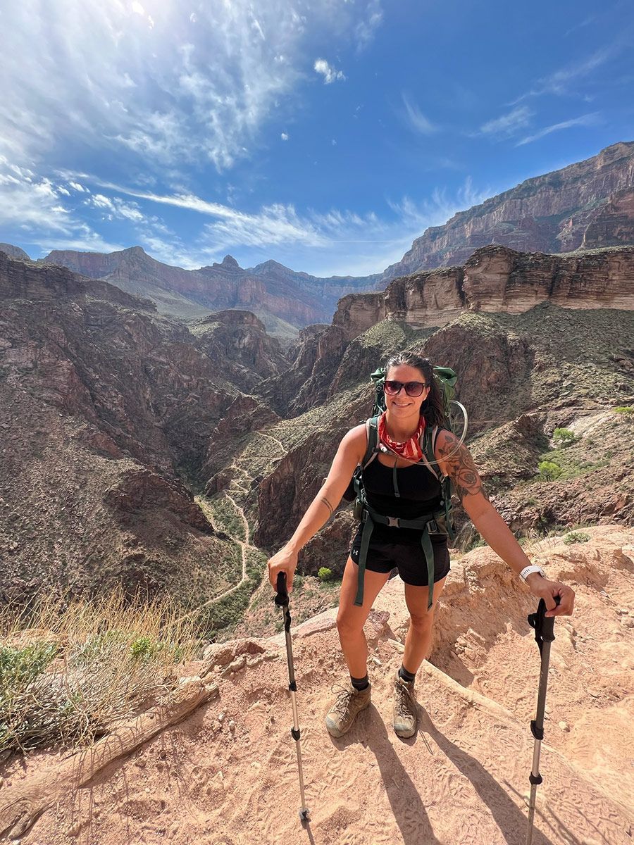 Woman hiking in a canyon, smiling at the camera, using hiking poles. Sunny day, rocky trail, mountain backdrop.
