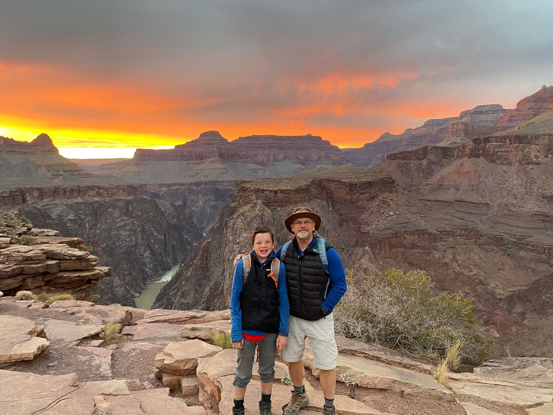 A man and a boy are standing on top of a canyon at sunset.
