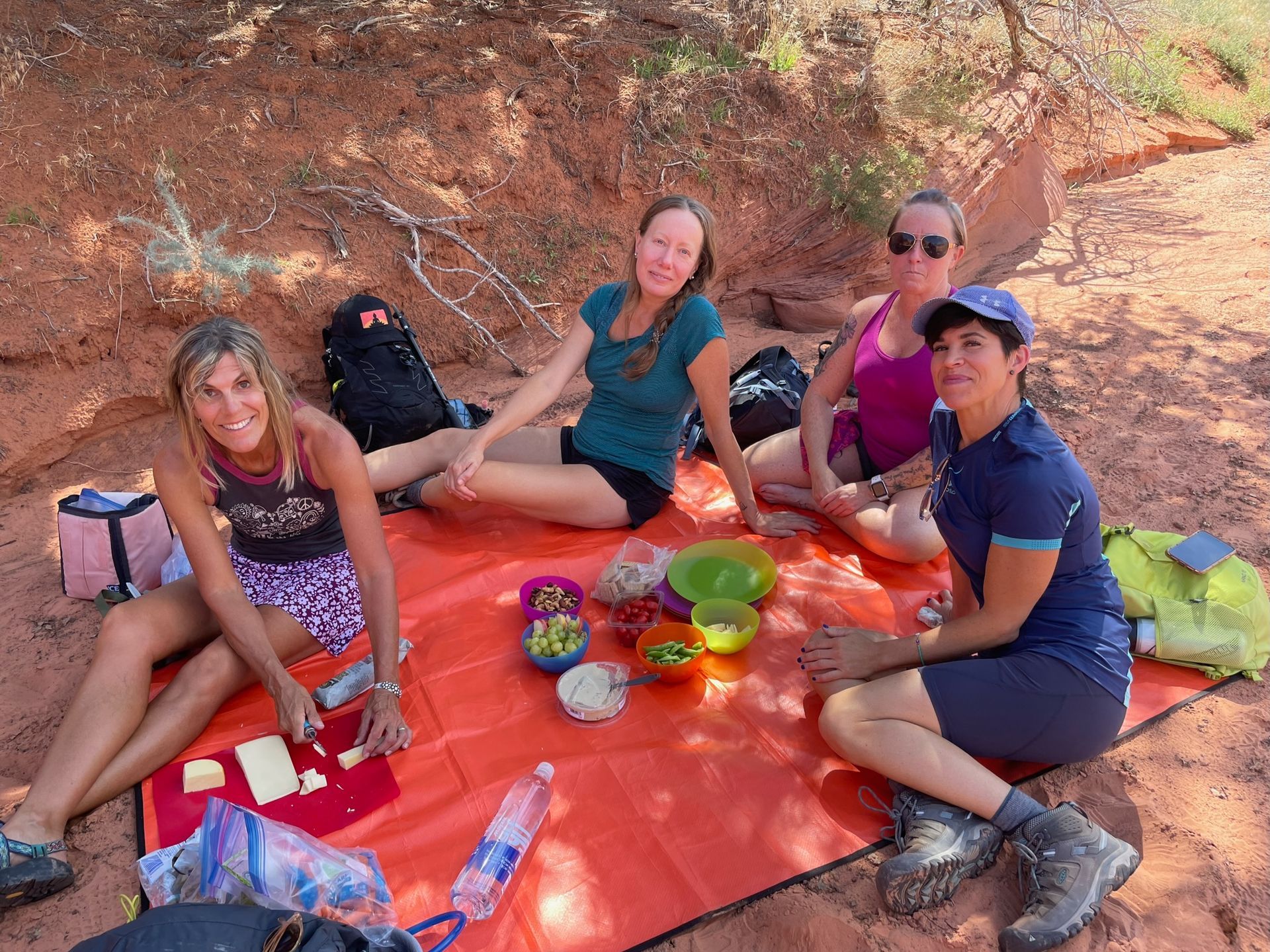 A group of women are sitting on an orange blanket in the dirt.