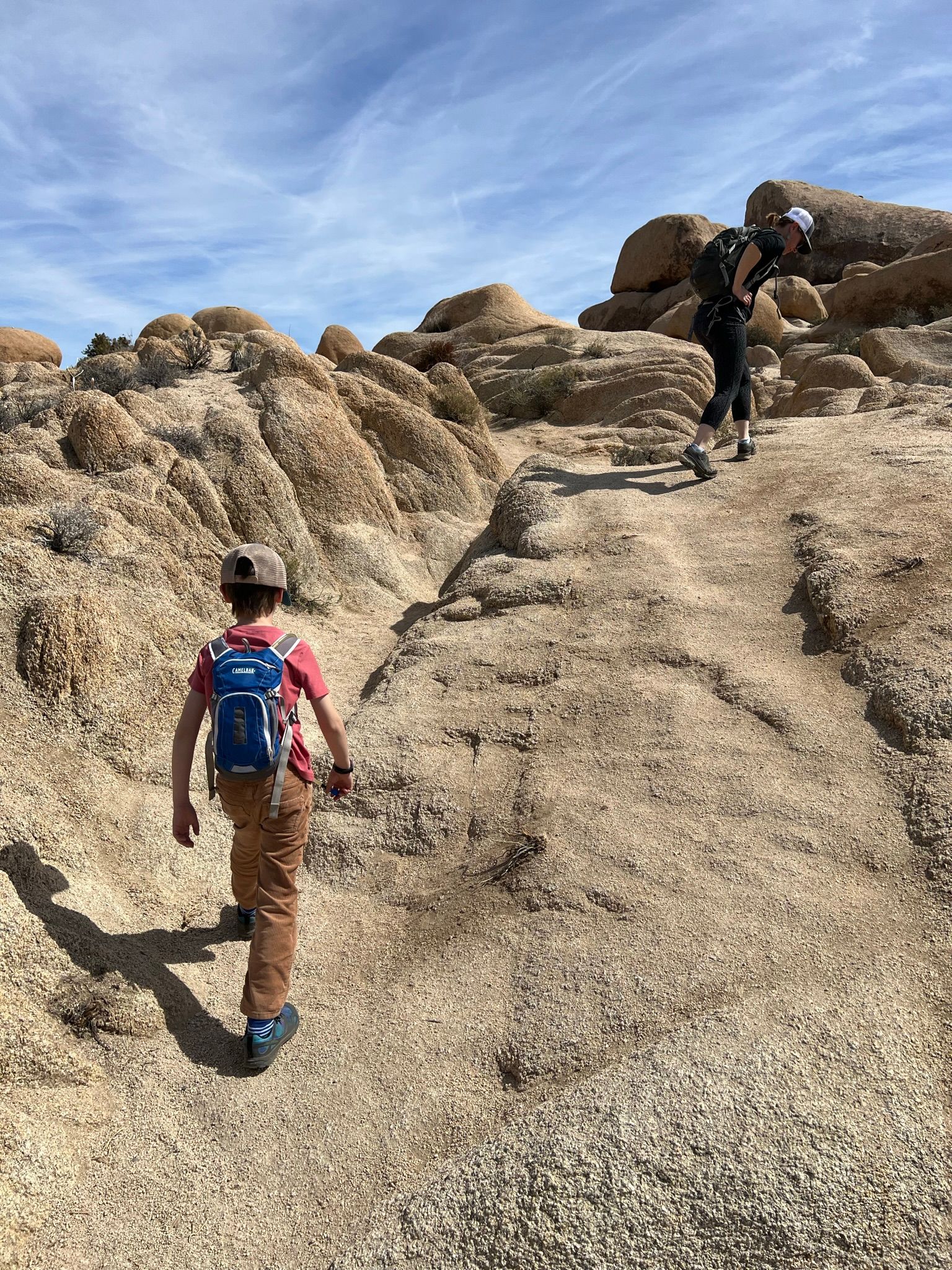 Two people hike up a rocky trail on a sunny day. Child in red shirt, boy in front, adult behind.