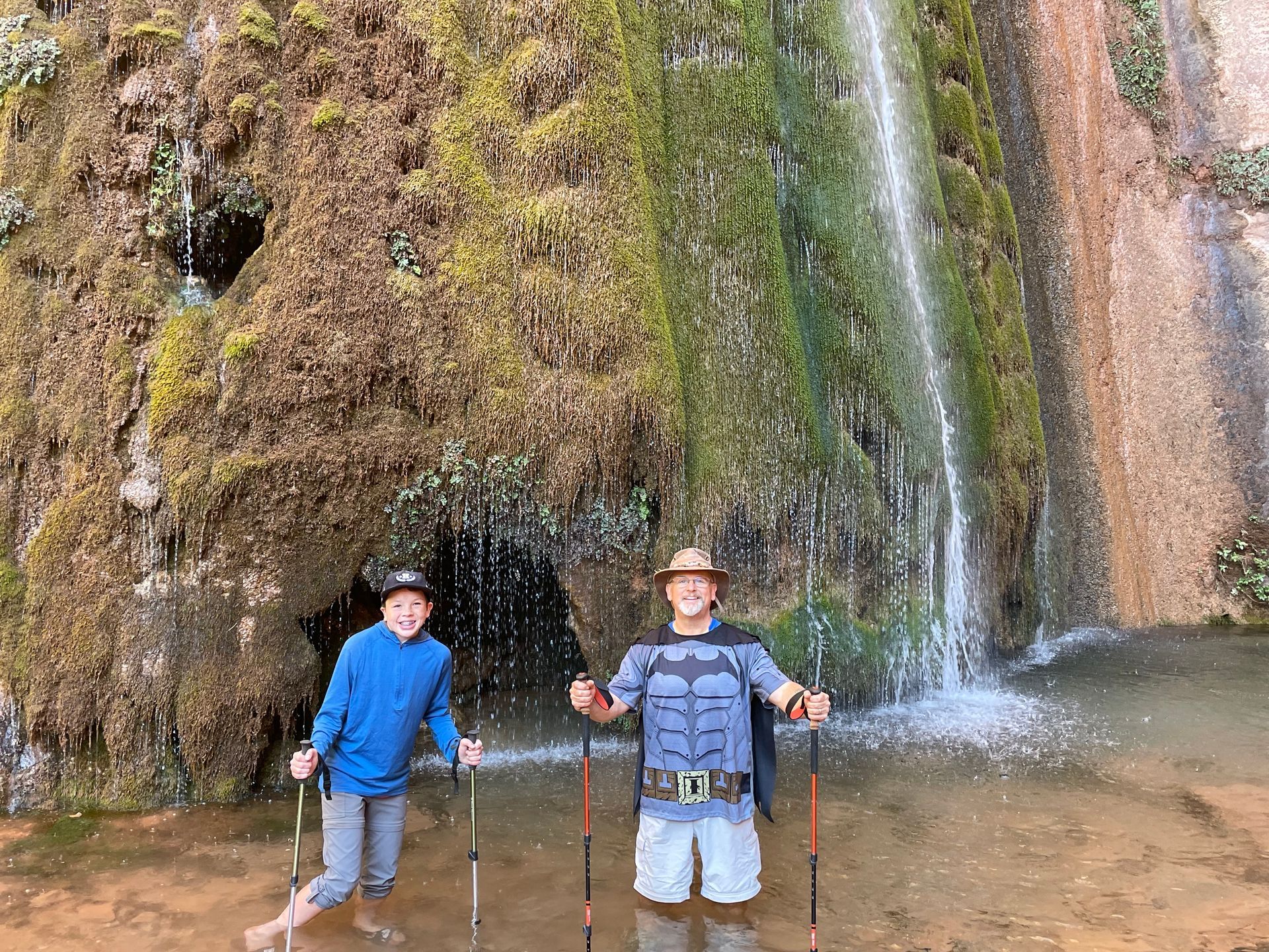 Two people are standing in front of a waterfall holding hiking poles.