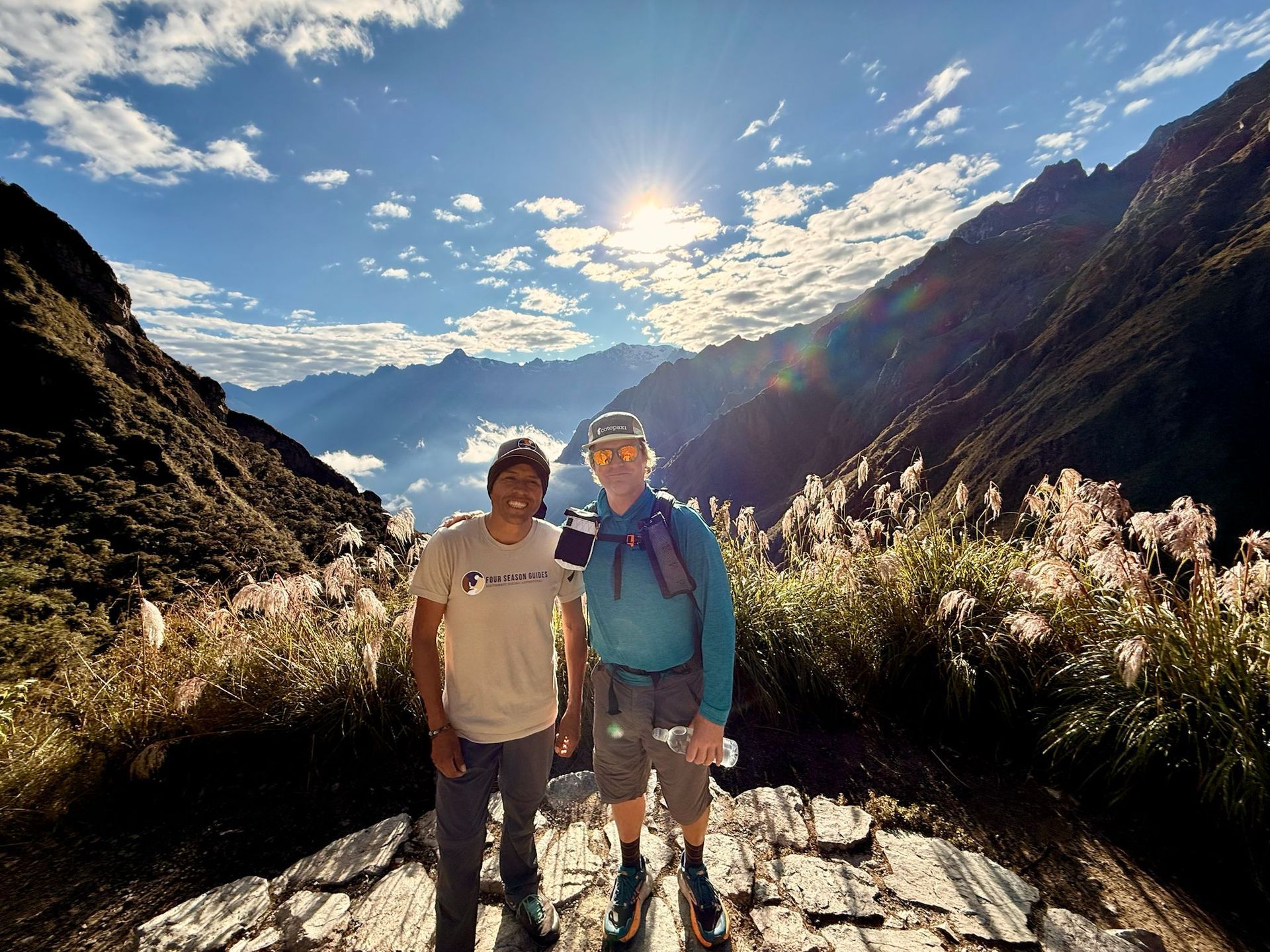 Man in shorts and jacket stands on a trail with mountain views.