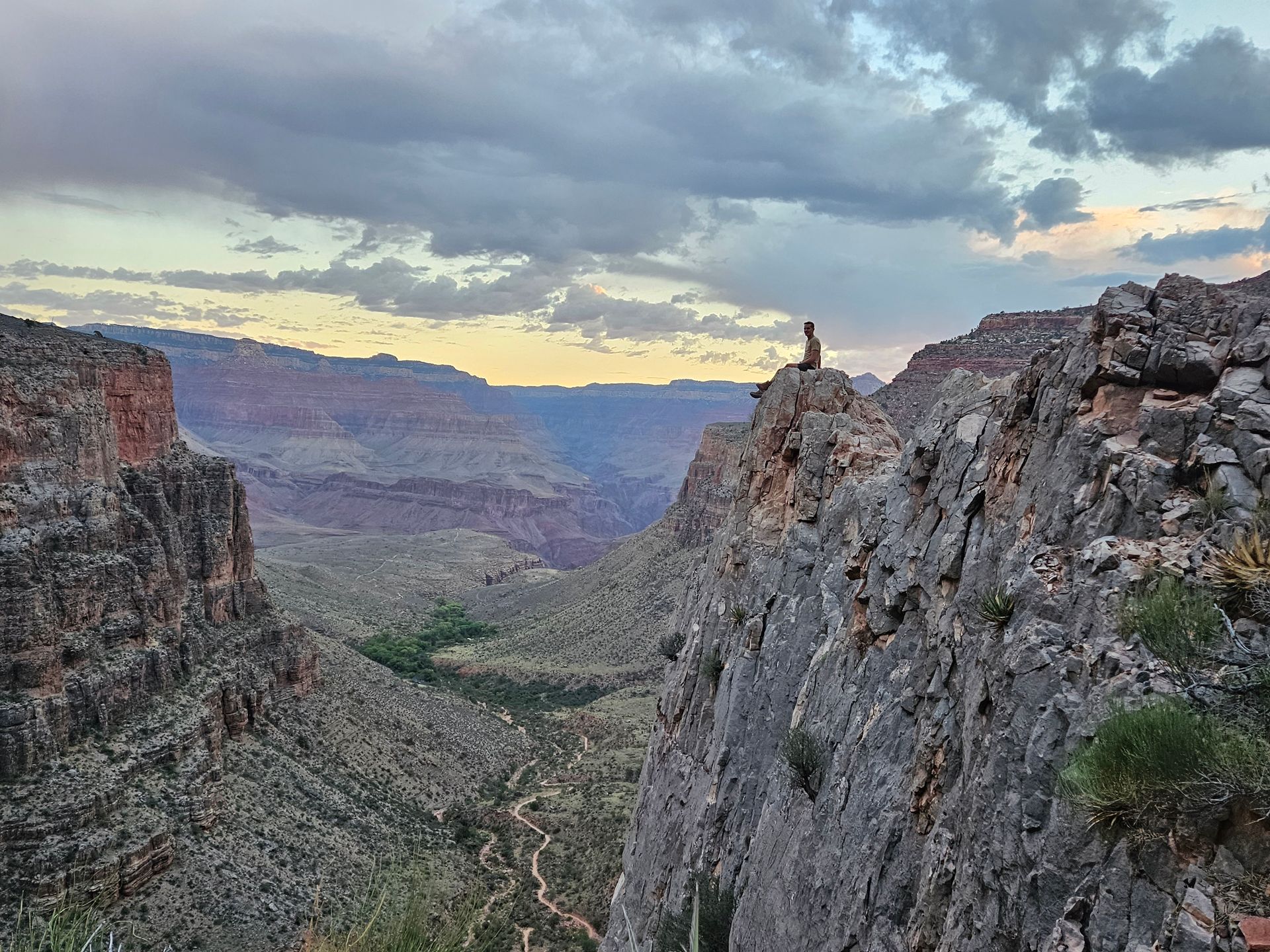 Person sitting on rocky cliff overlooking canyon at sunset.