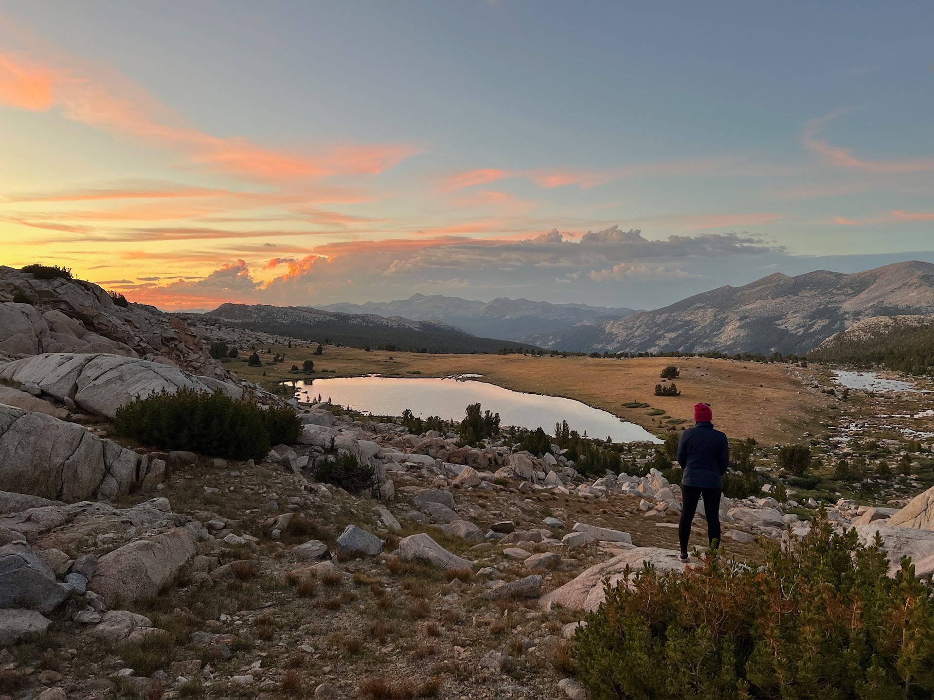Person overlooking a mountain lake at sunset, pink clouds, rocky terrain.