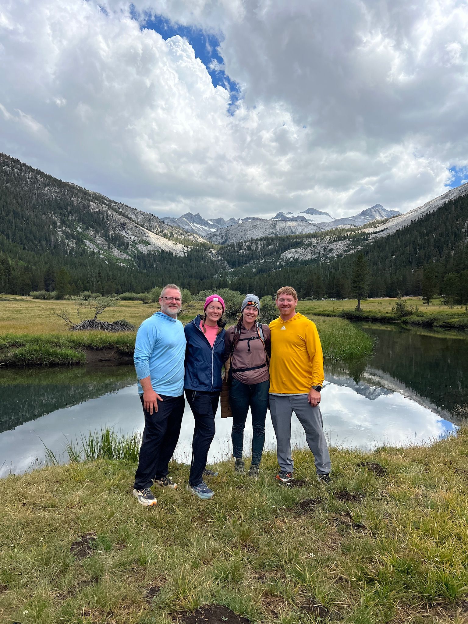 Four people stand near a lake, mountains in background, cloudy sky.