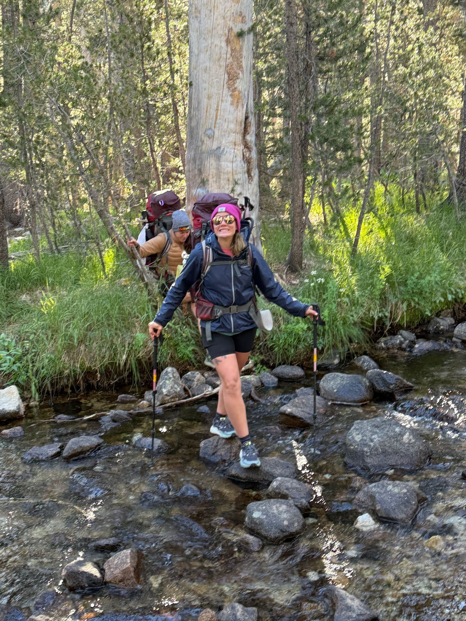 Woman hiking in stream, using poles for balance, another hiker behind in forest.