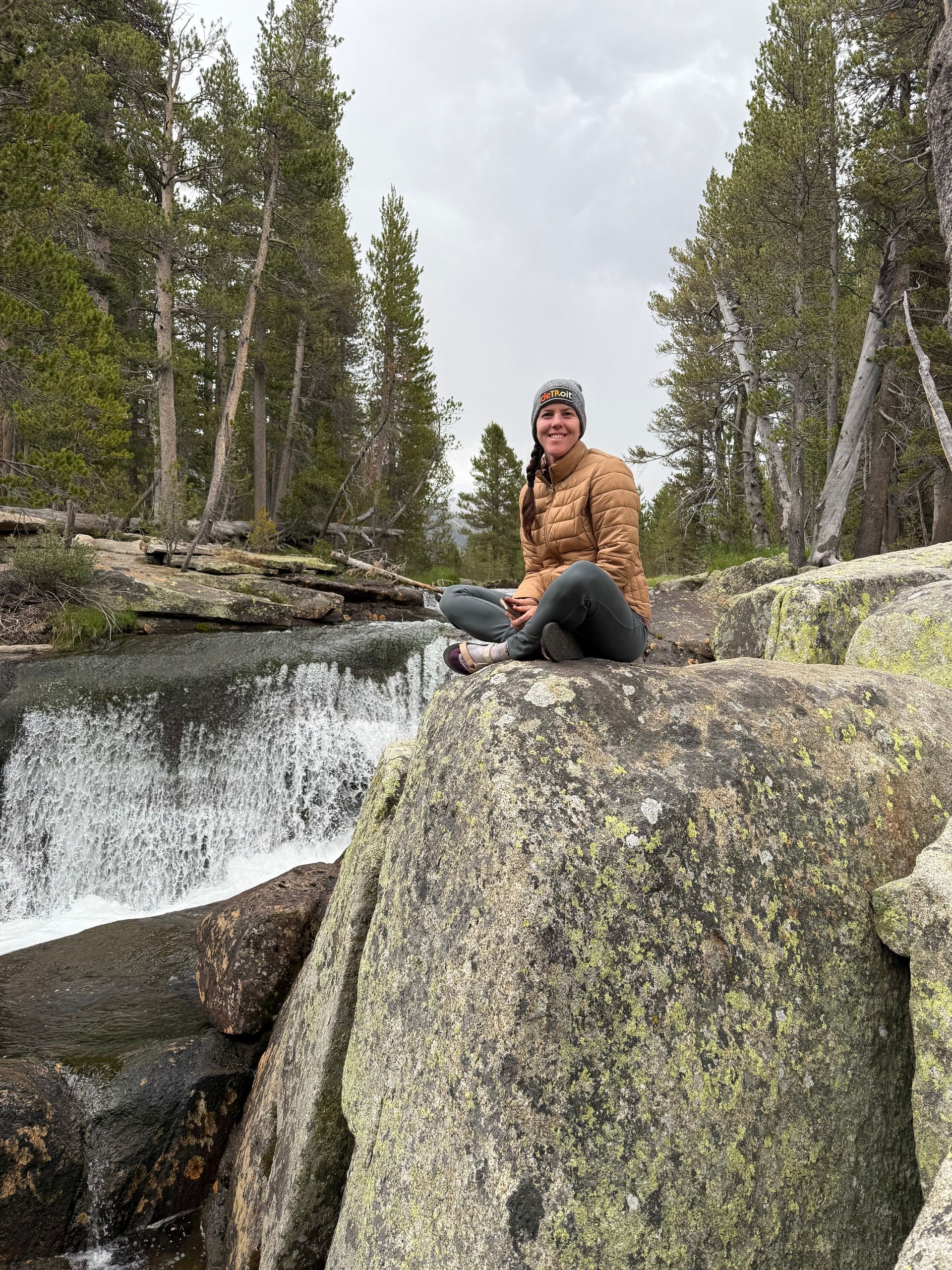 Person sits on a rock near a waterfall in a forest. They wear a jacket and hat.