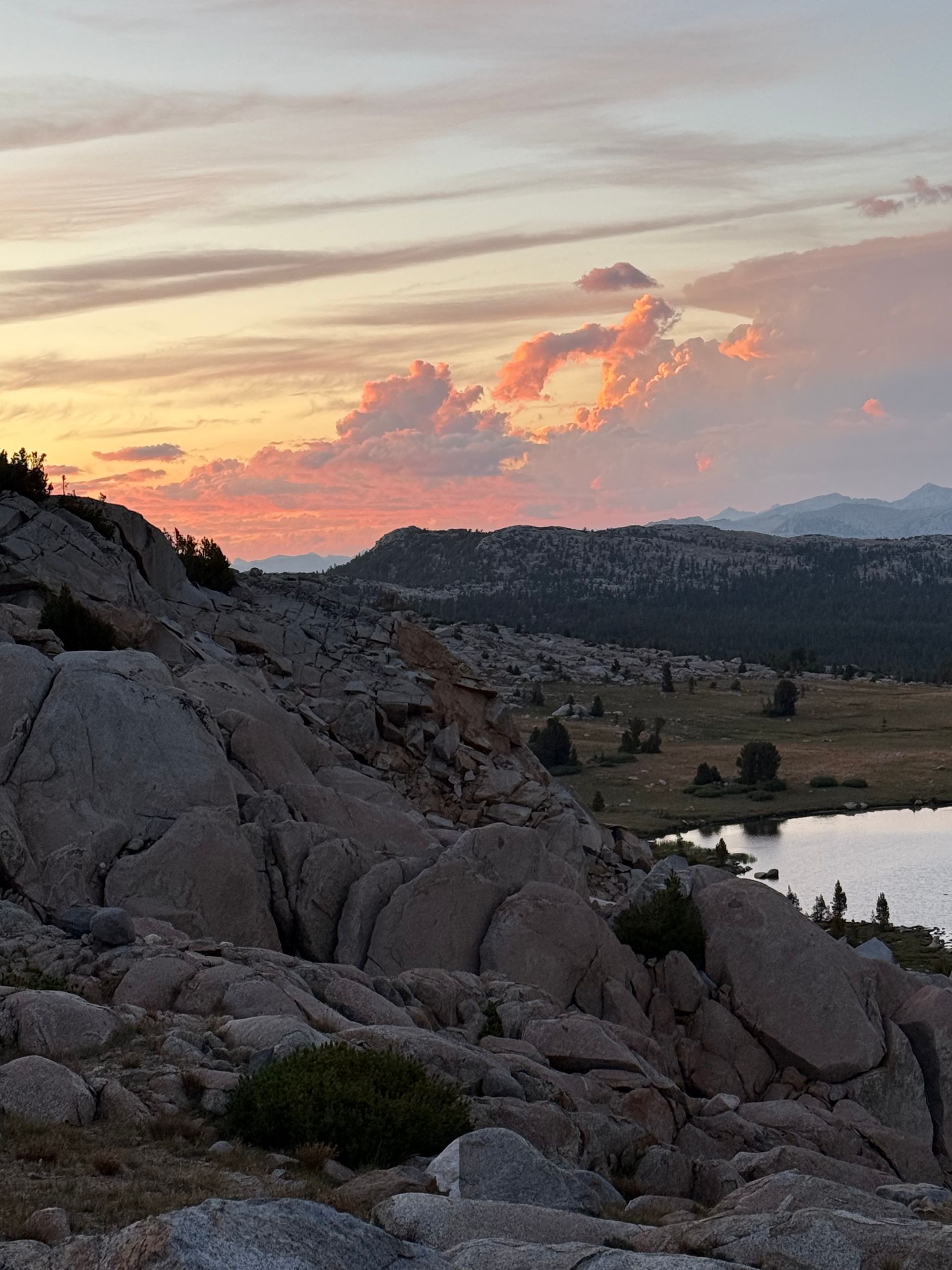 Rocky mountain landscape at sunset, pink and orange clouds, lake, and trees.