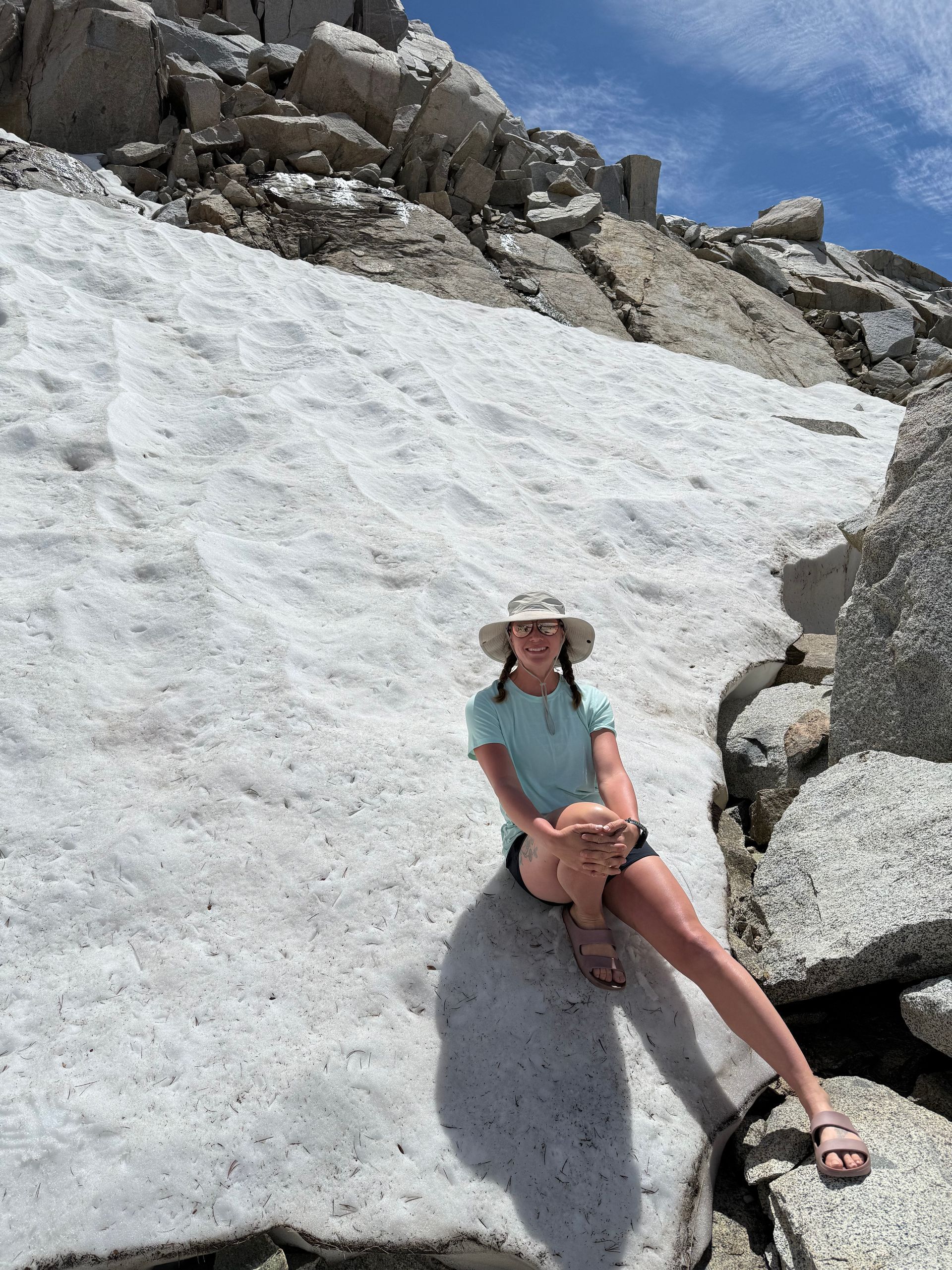 Person sitting on snow near rocks, wearing a hat and teal shirt. Sunny mountain setting.