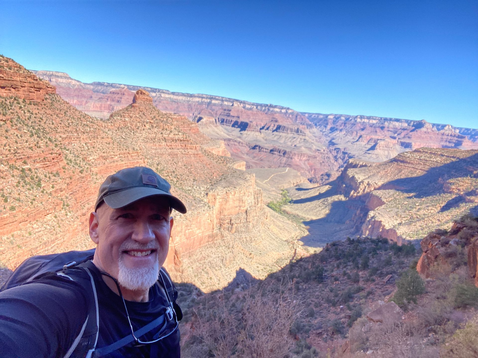 Man with gray beard smiles, poses for selfie in front of the Grand Canyon on a sunny day.