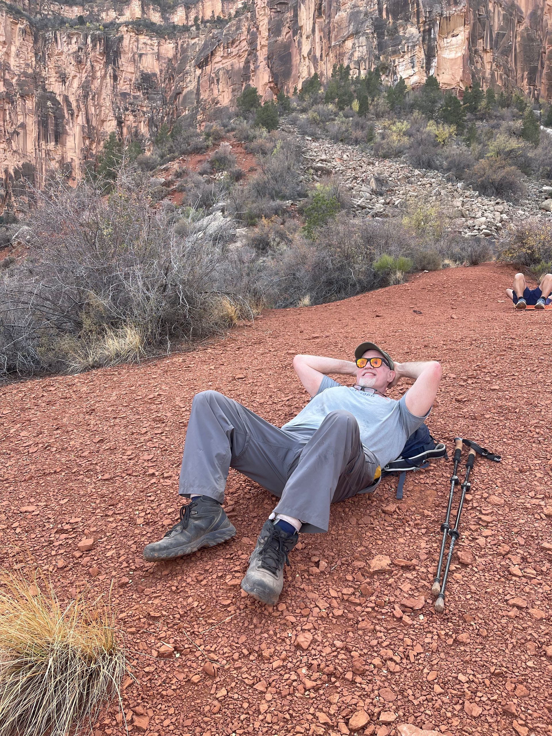 Man resting on red dirt, arms behind his head, hiking boots, canyon backdrop.