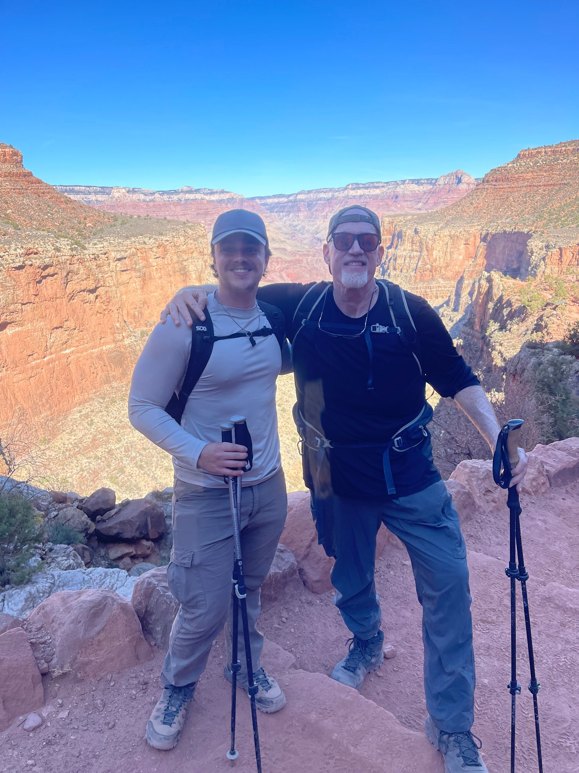 Two hikers pose with arms around each other on a red rock trail overlooking a canyon.