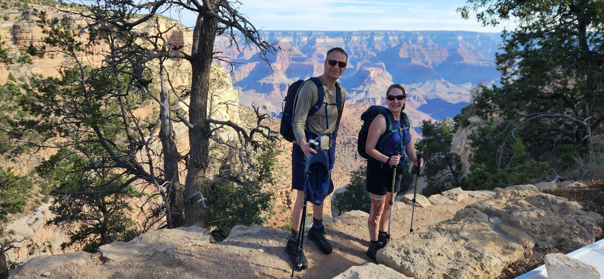 Two hikers pose on a cliff edge overlooking the Grand Canyon. They wear backpacks and sunglasses; trees frame the view.
