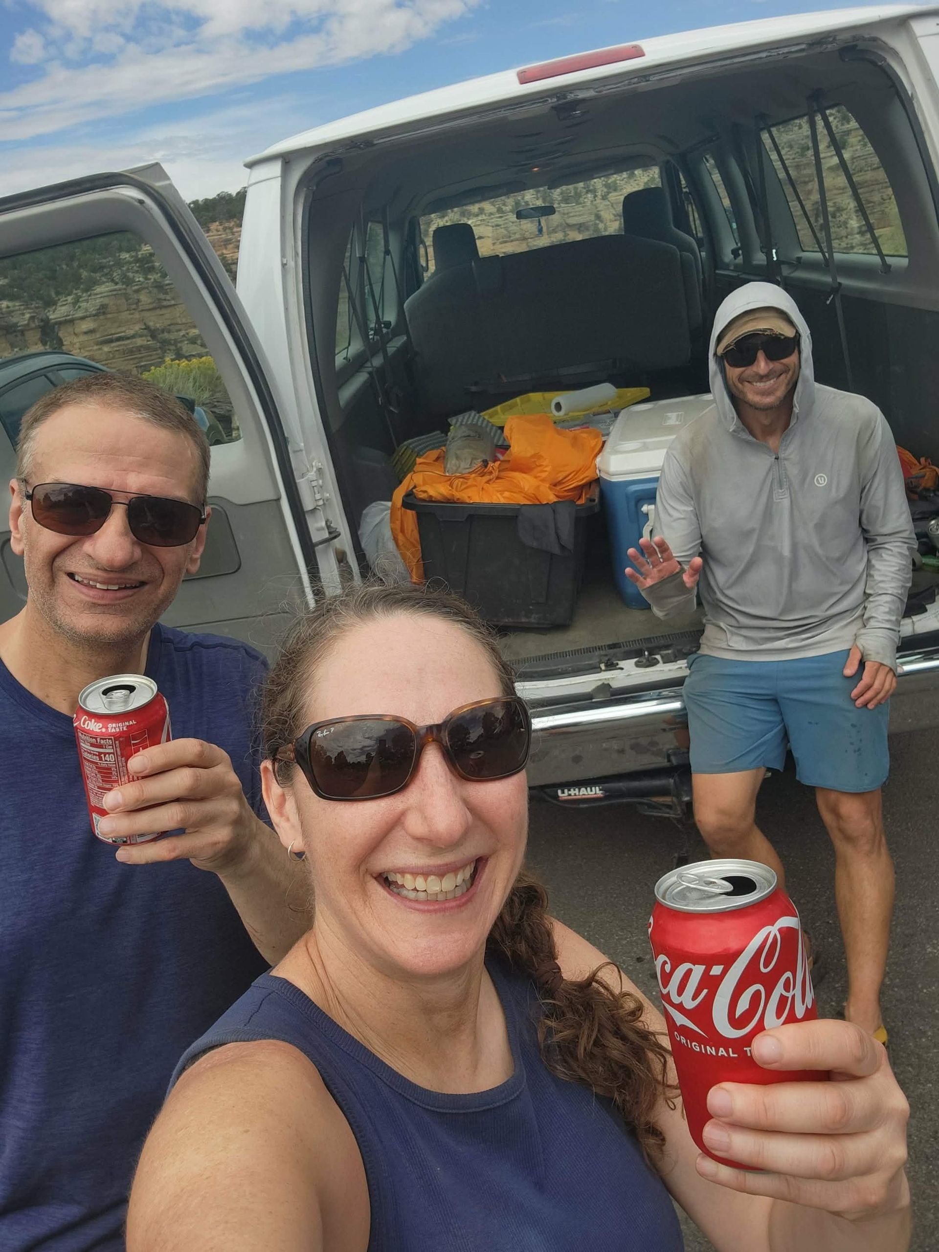 Three people holding Coca-Cola cans, smiling near an open van. Sunny outdoor setting.