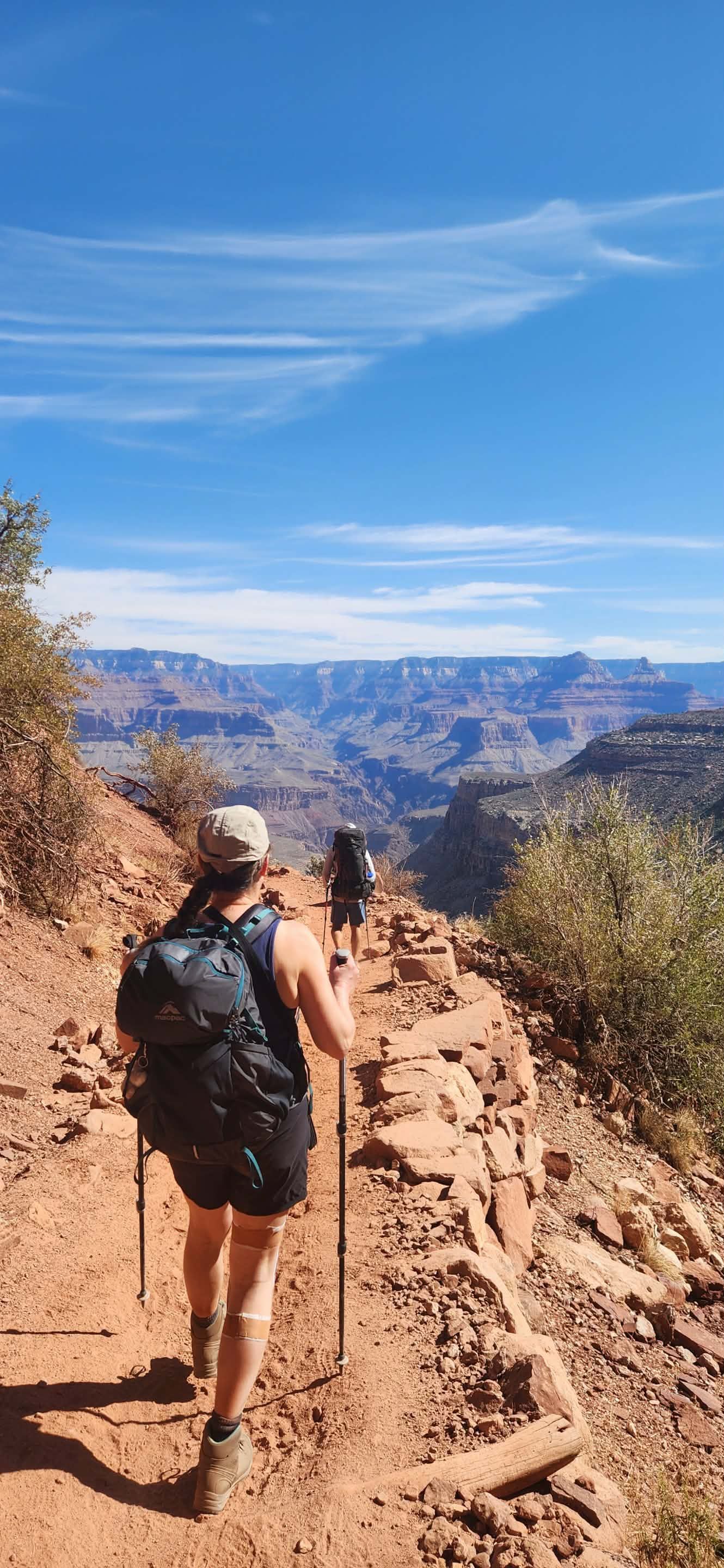 Hiker walking on a red dirt trail with canyon views under a blue sky.