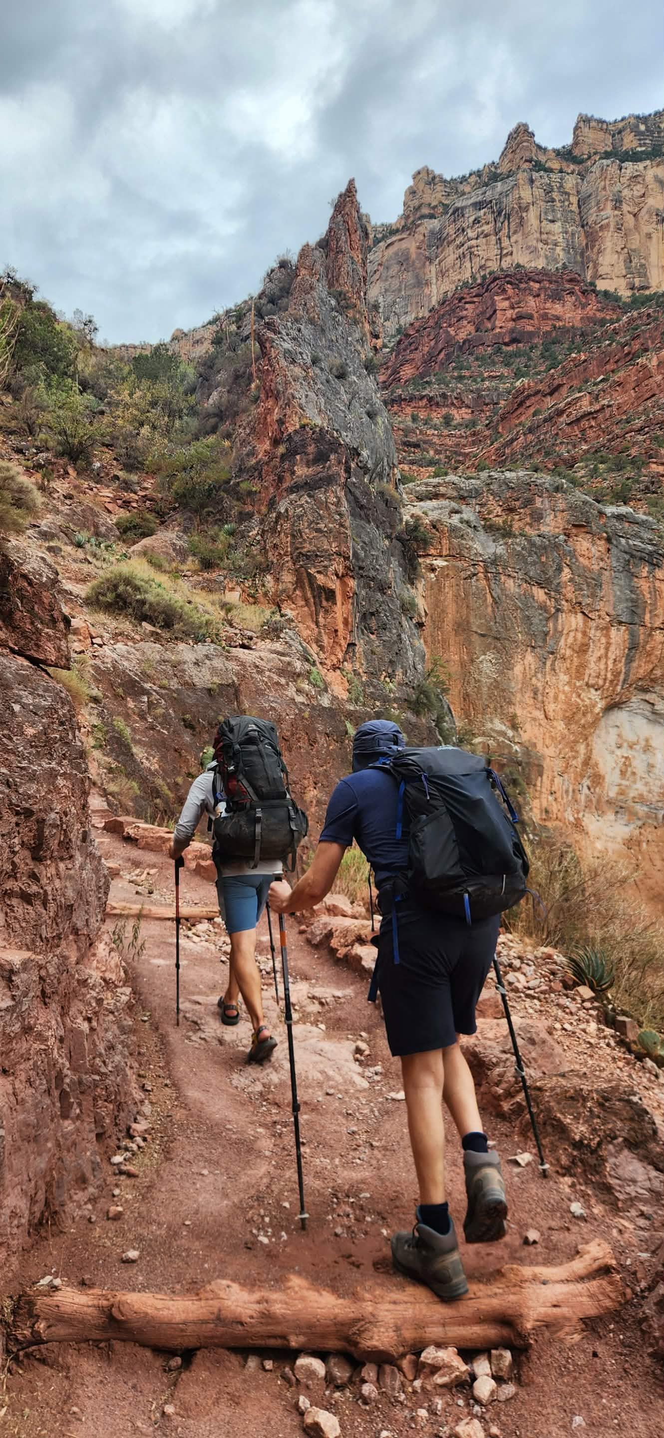 Two hikers ascend a rocky trail in a canyon, using trekking poles. Red rock cliffs and overcast sky.